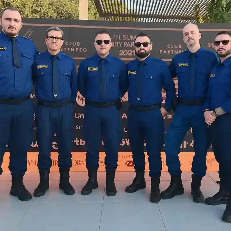 Sei uomini in uniforme blu e occhiali da sole posano davanti allo sfondo di un evento in discoteca.