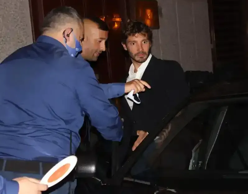 Di notte, due uomini con giacche scure sono vicino a un'auto: uno indica qualcosa mentre l'altro si sporge verso la portiera aperta.