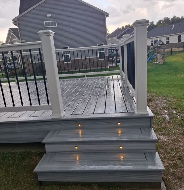 Gray deck with steps lit by small lights; black railing. Houses and grass in background.