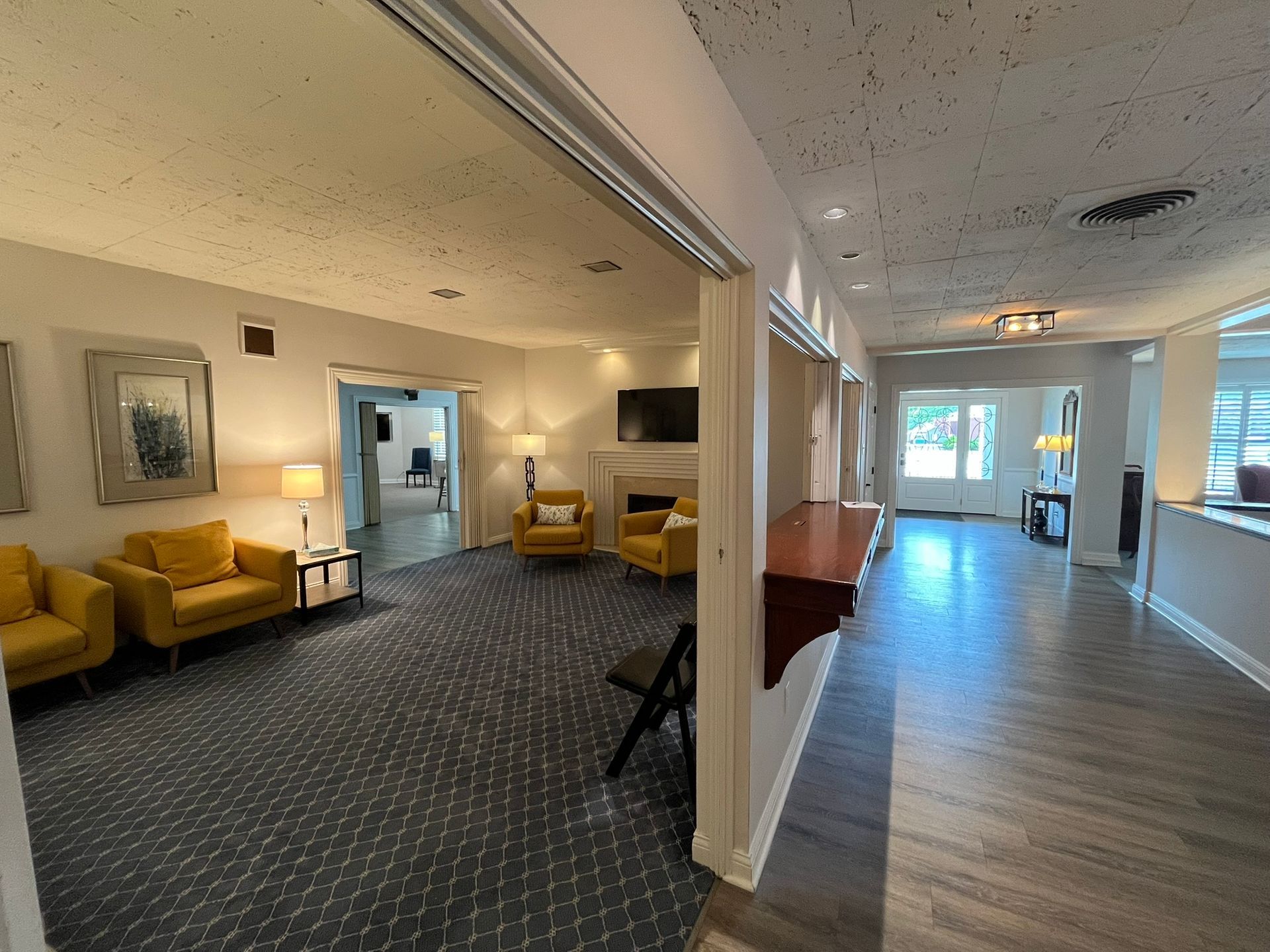 Bright lounge with yellow chairs, patterned carpet, and wood flooring leading to a windowed hallway