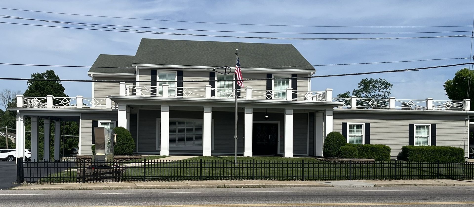 White-columned two-story building with a porch and flags behind a black fence under power lines