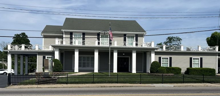 White-columned two-story building with a porch and flags behind a black fence under power lines