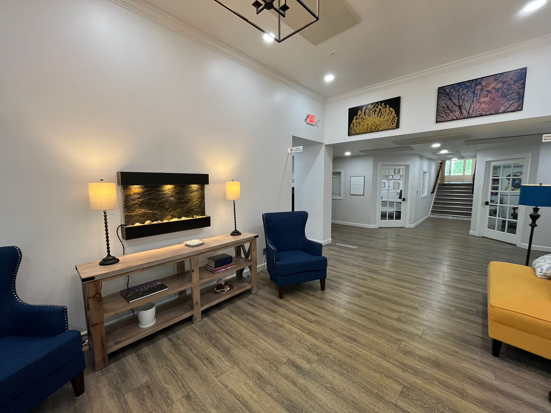 Bright lobby with wood floors, fireplace TV, blue chairs, yellow bench, and framed art above a hallway.