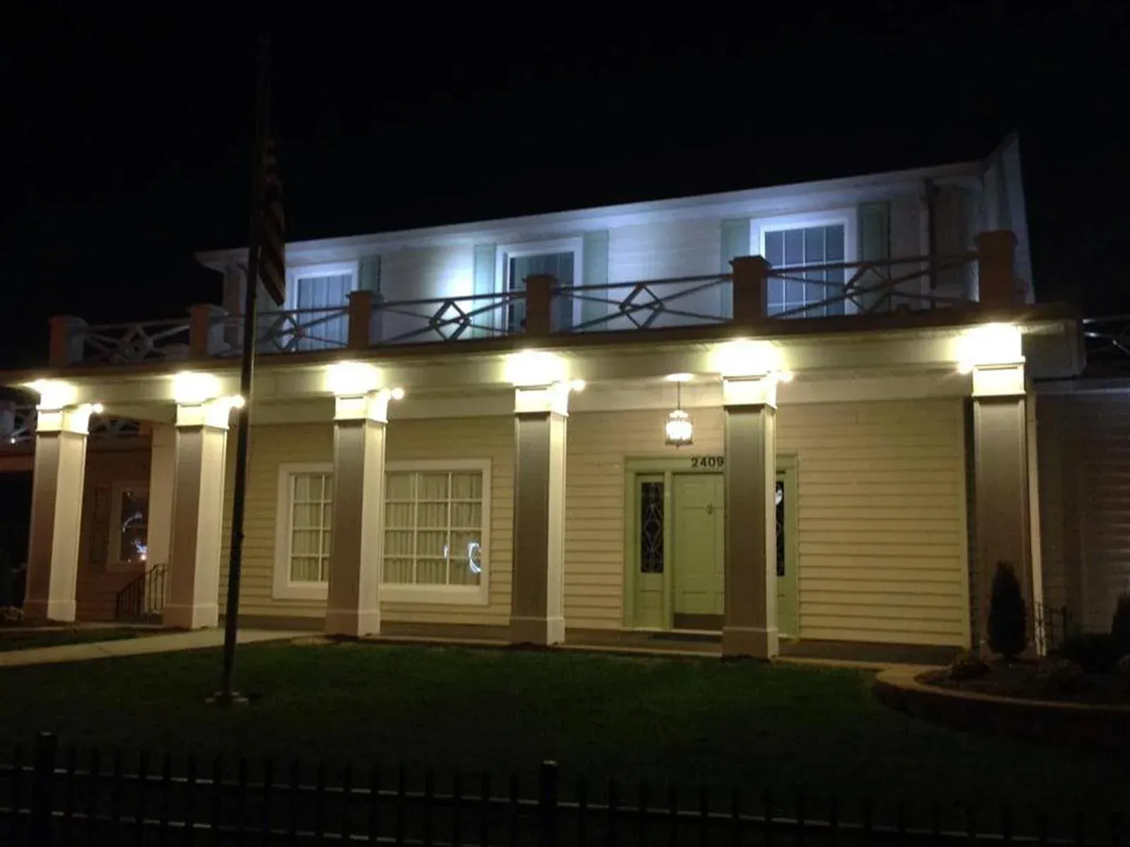 Well-lit two-story building at night with white columns, balcony railing, and green door and lawn