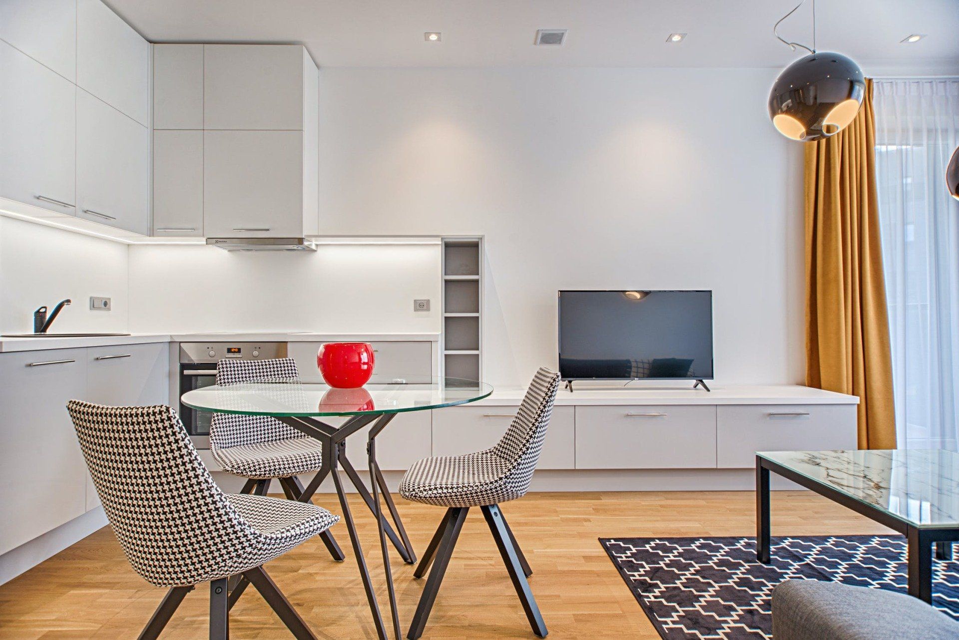 Modern, bright studio apartment with kitchen, dining area, and TV. Features white walls, wooden floors, and patterned chairs.