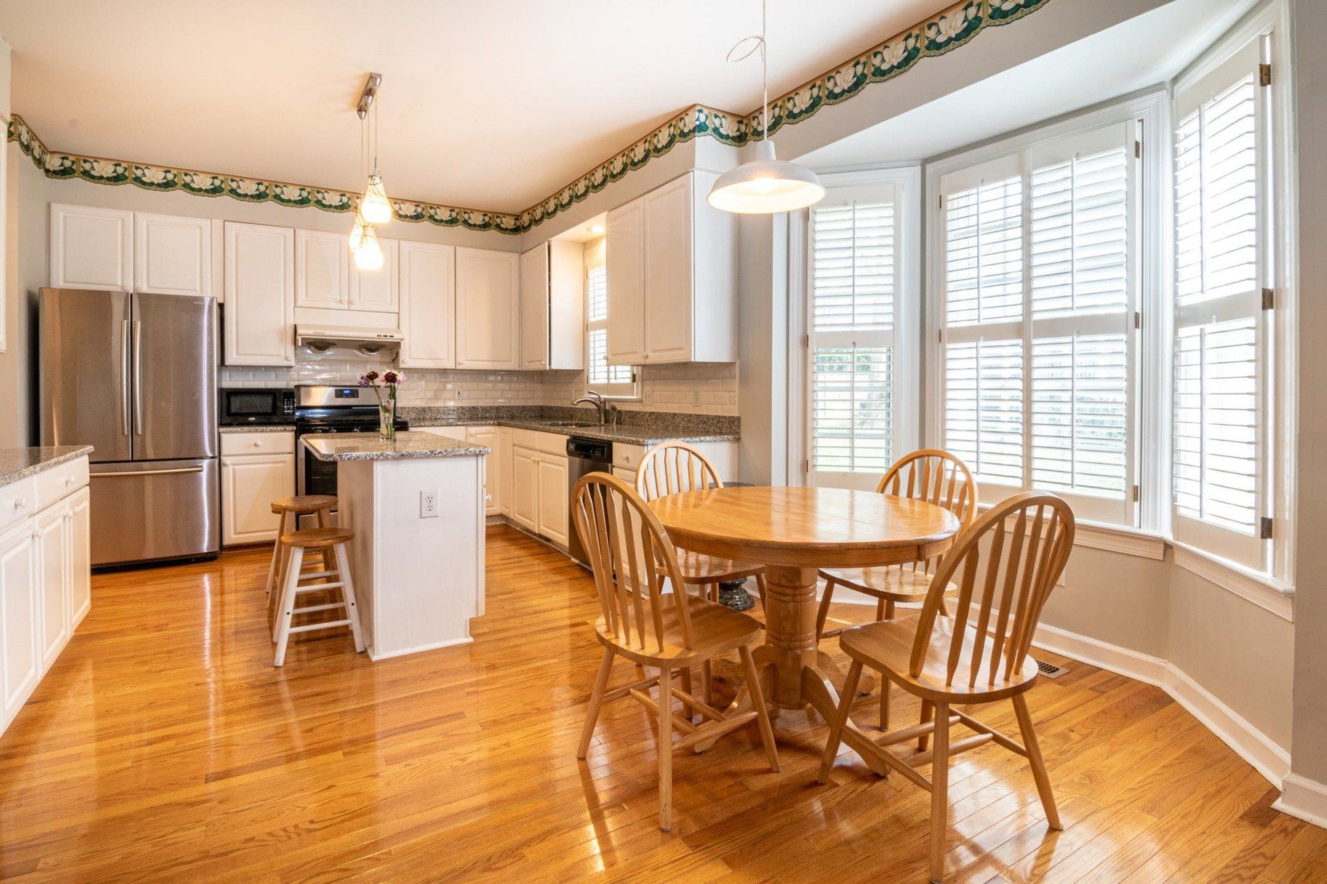 Kitchen with white cabinets, stainless steel fridge, island, and dining table by a window with shutters.