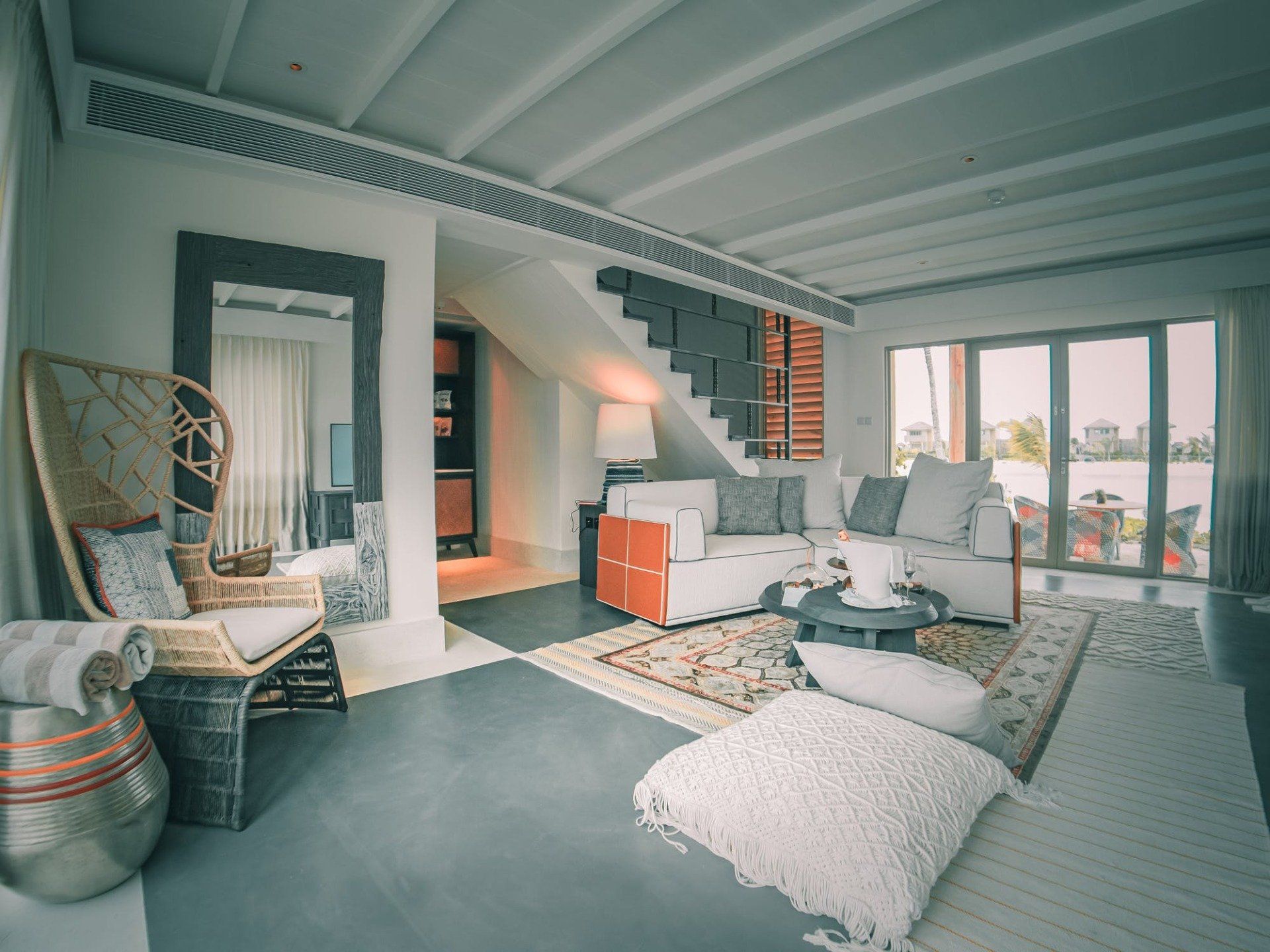 Bright living room with stairs, wicker chair, sofa, rug, and large windows.