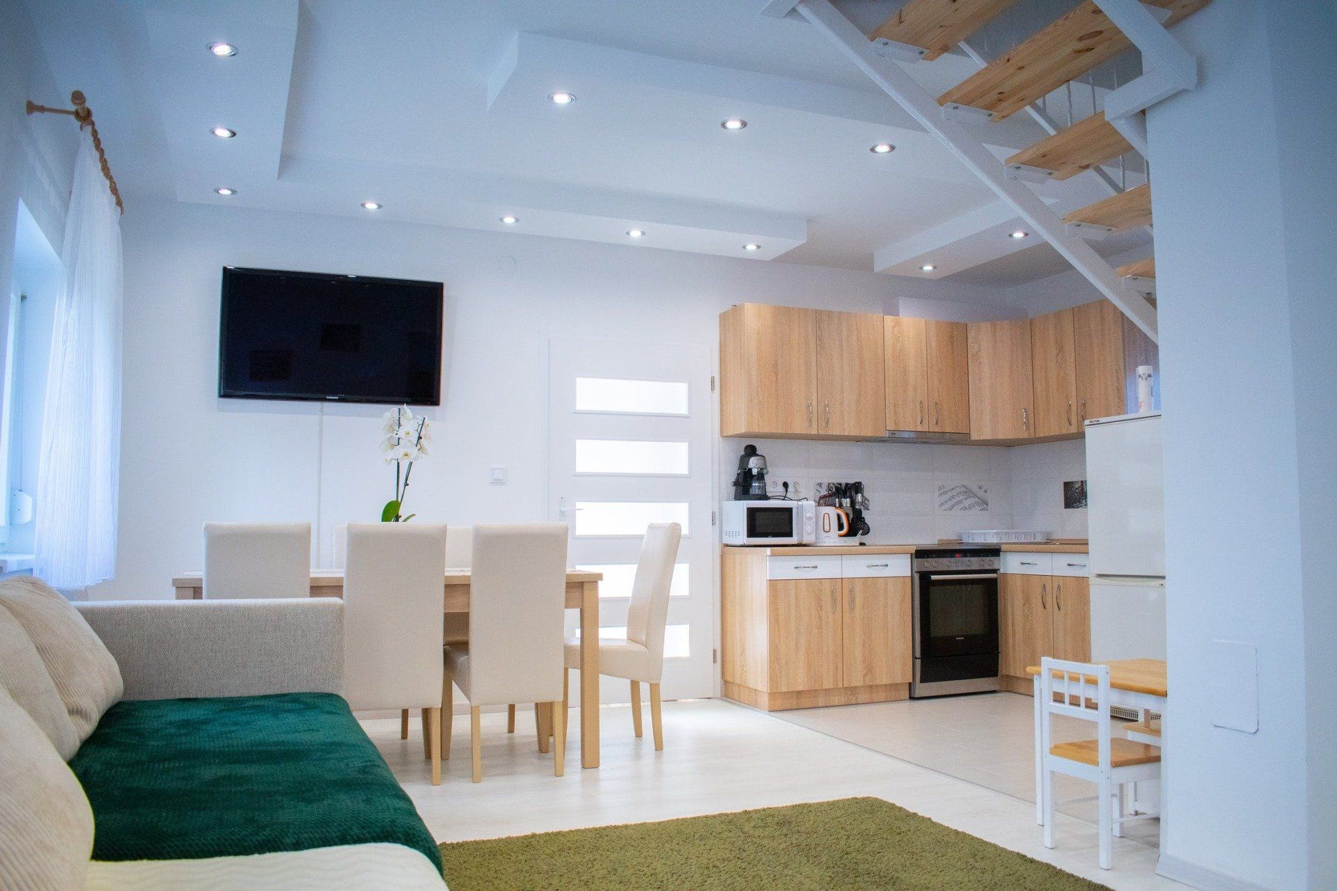 Living space with kitchen, dining area, and stairs. White walls, wooden cabinets, and green accents.