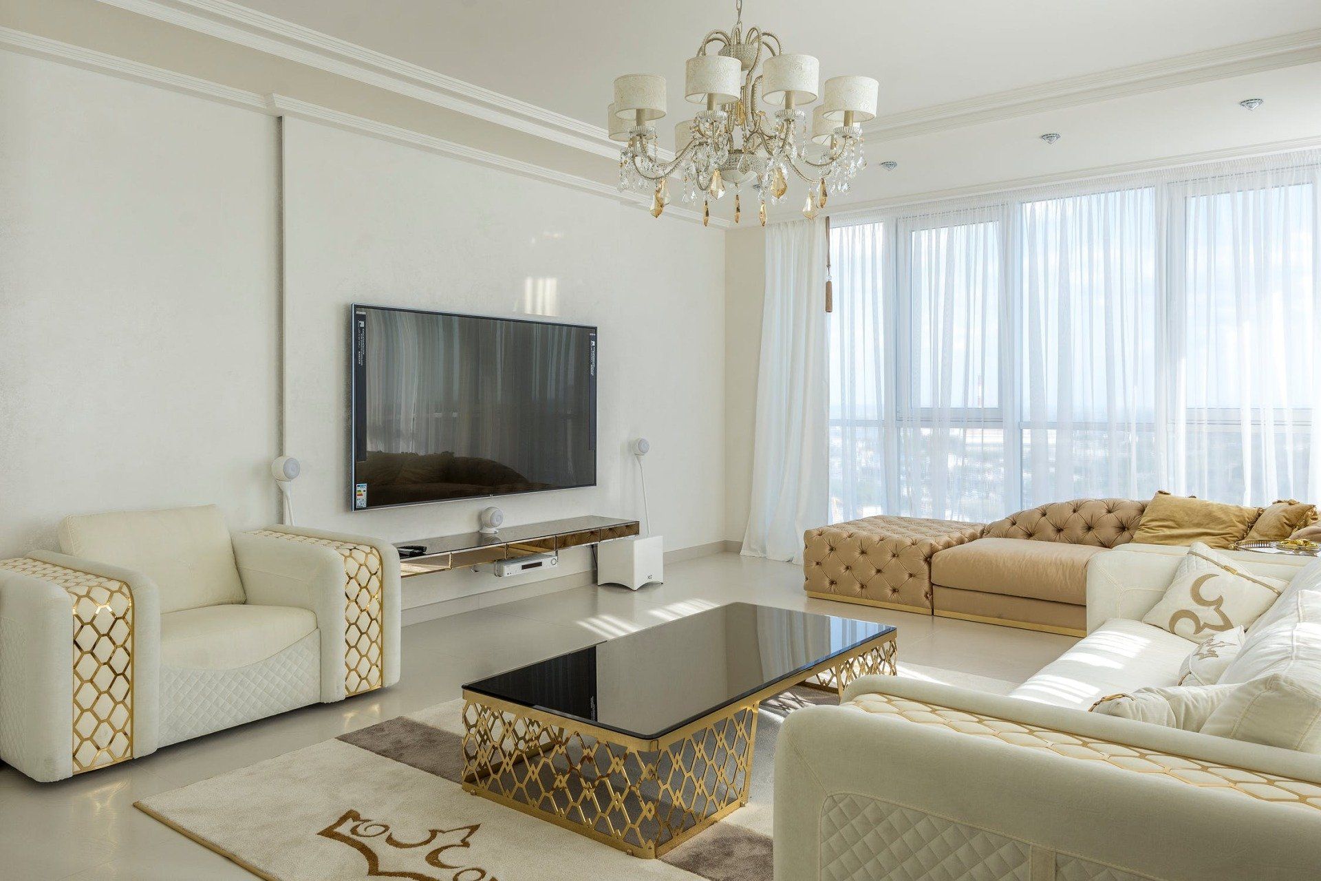 Luxurious living room: white furniture, gold accents, large TV, chandelier, and a large window with white curtains.