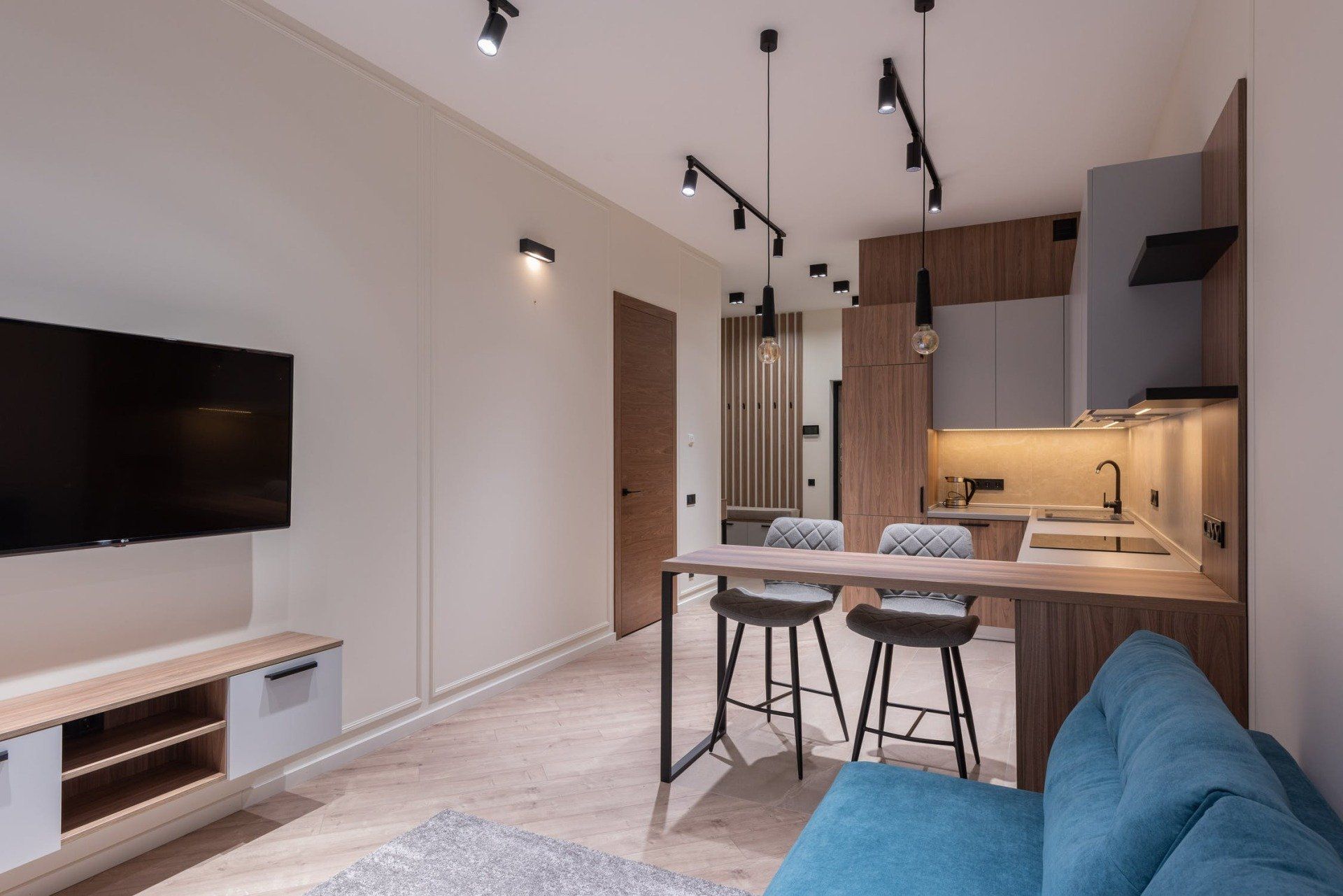 Modern, open-concept living space with kitchen and living area. Features teal sofa, wooden cabinets, and bar seating.