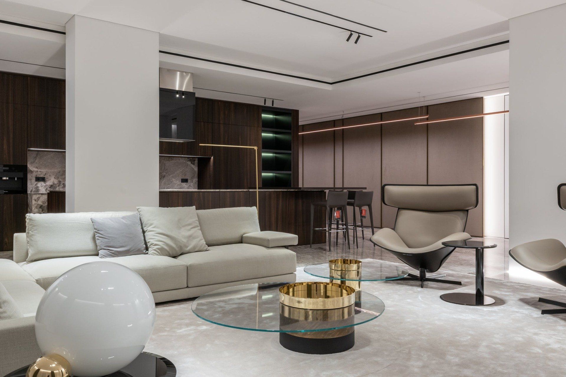 Modern living room with kitchen. Beige sofa, glass table, and gray accent chair. Dark wood cabinetry.