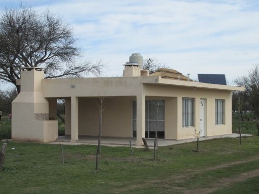 Pequeña casa de color canela con patio cubierto, parrilla al aire libre y tanque de agua, ubicada en un jardín con césped.