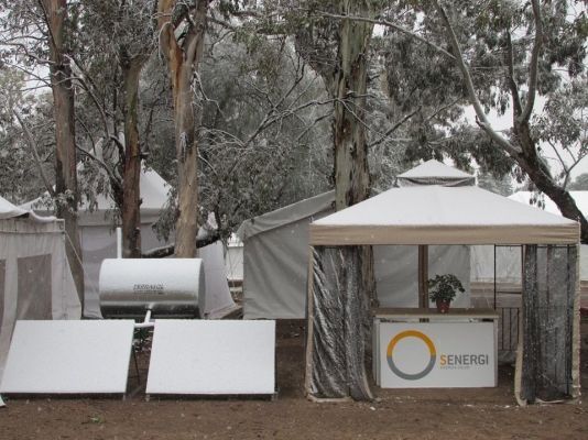 Escena nevada: stand de Generac con paneles solares y tanque de agua, árboles al fondo.