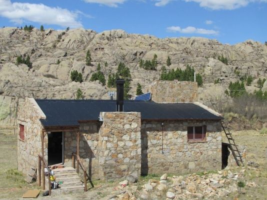 Cabaña de piedra con techo negro, chimenea y escalera contra una ladera rocosa bajo un cielo azul.