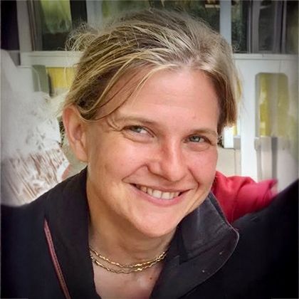 Closeup photo of smiling white woman's face. She wears red framed glasses and looks straight at the camera. Her hair is white. She seems to be sitting in a chair outdoors in sunshine, but the photo is so closely cropped it's hard to tell.