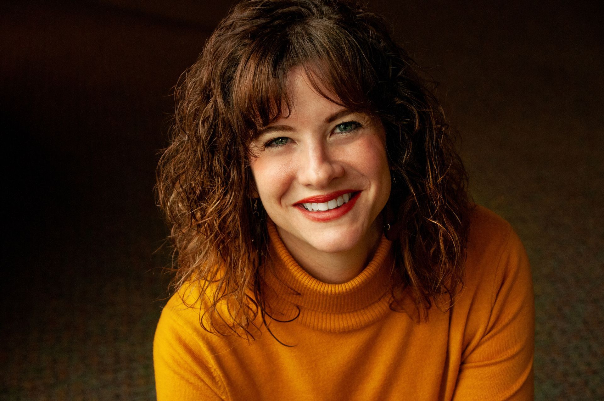 Smiling white woman with brown wavy hair and blue eyes, wearing a harvest gold turtleneck sweater.