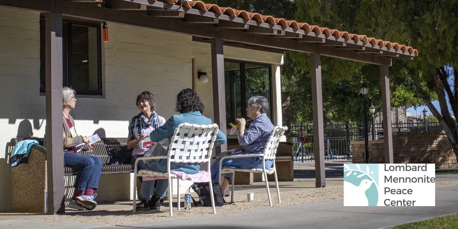 A photo shows a group of ladies in casual clothes sitting together in a miscellany of chairs under the roof of a Spanish style building, eating and talking on a sunny day.  The Lombard Mennonite Peace center logo is visible on the bottom right.