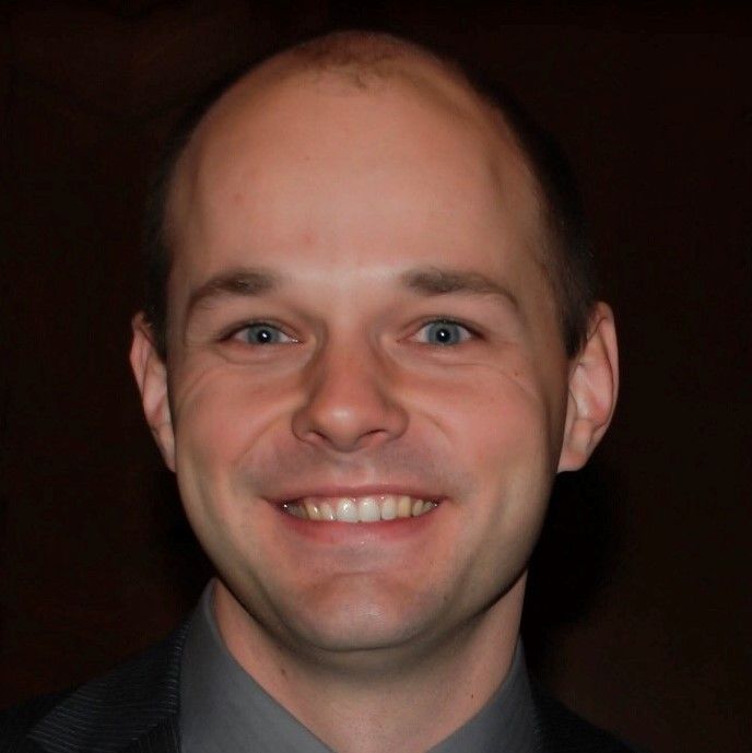 Closeup photo of a smiling white man's face against a dark background. He has short dark hair and grey eyes.