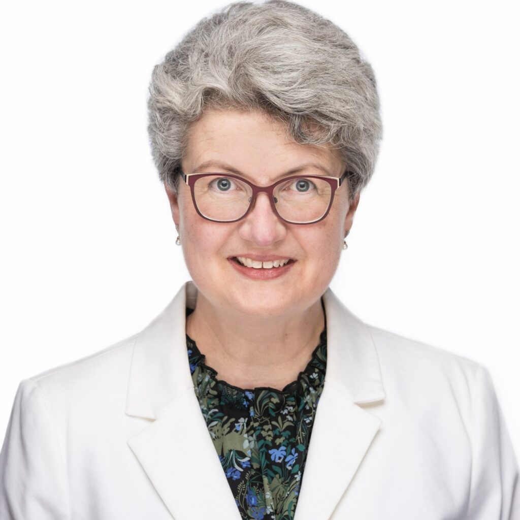 Mugshot of smiling white woman with short grey hair, red glass frames, a white blazer and floral black blouse. The background is white.