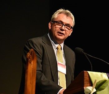 Photo of white man standing at a pulpit, his mouth open speaking. He wears a dark suit, white shirt and yellow tie. He has dark glass frames and white hair. He stands against a dark background, leaning into the pulip microphone.