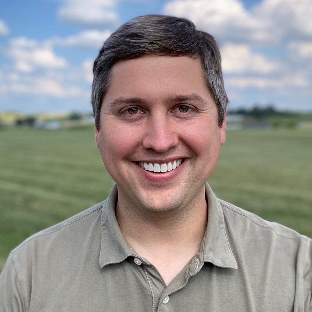 Mugshot of smiling white man in grey shirt with green fields and blue skies visible over his shoulder. He has dark brown hair with touches of grey, and looks straight at the camera.