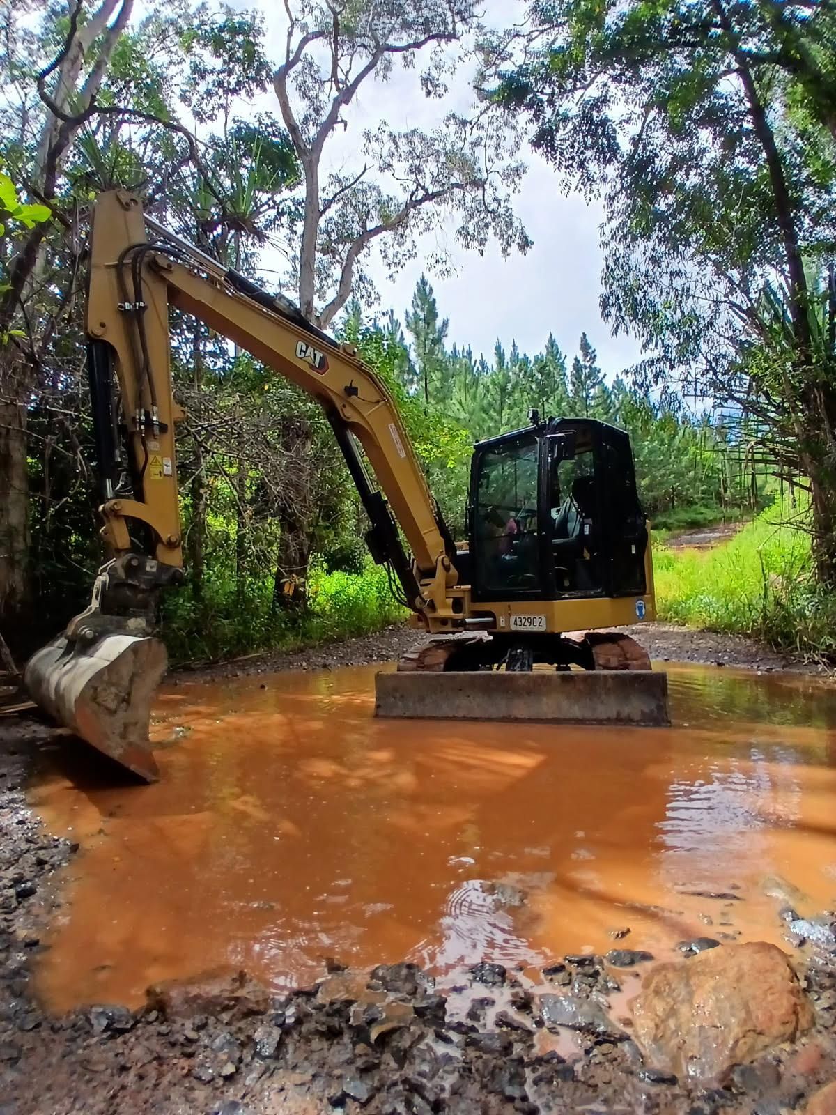 Yellow excavator in muddy water, surrounded by trees — Kuranda Landscape Supplies in Kuranda, QLD