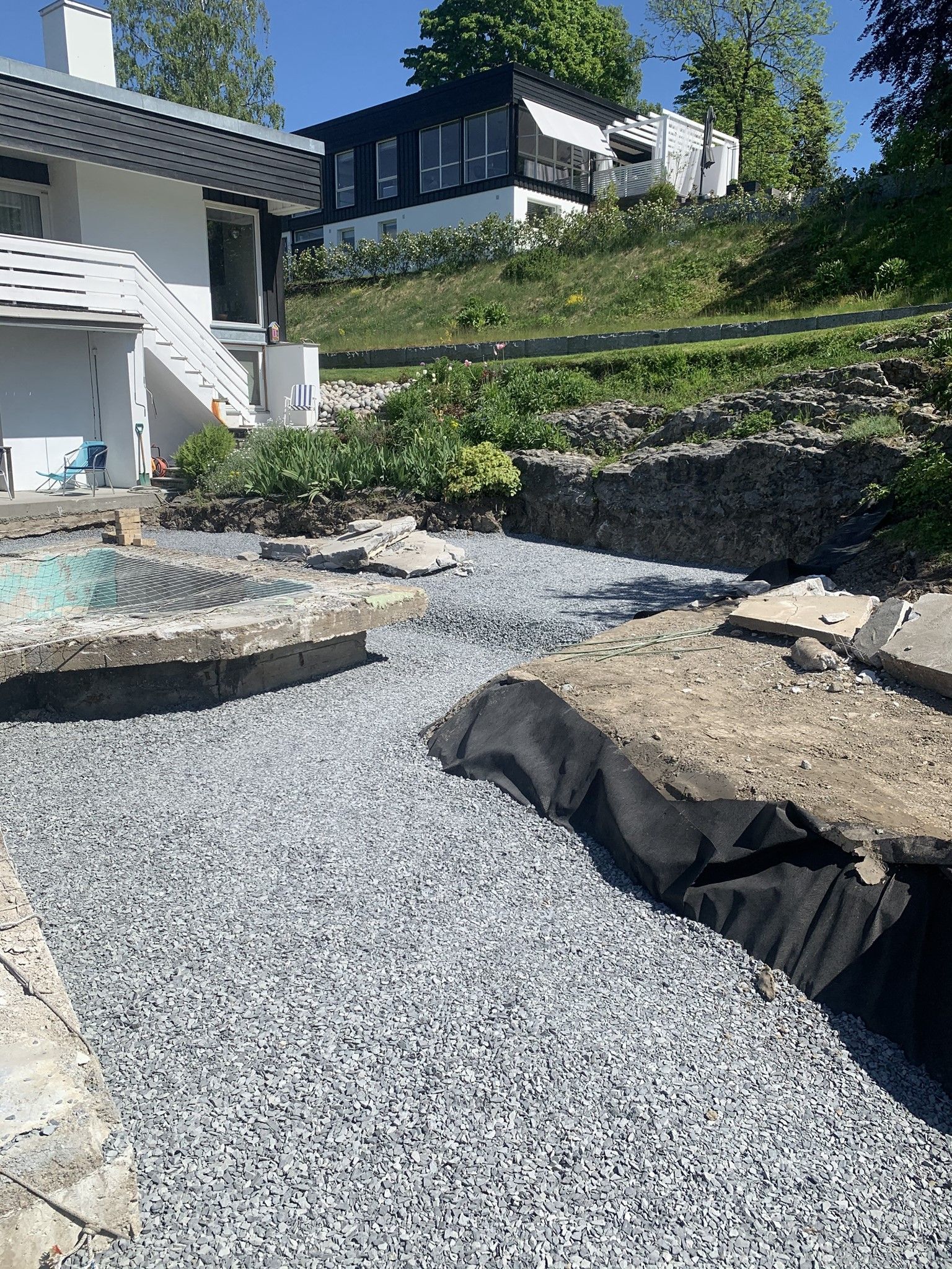 Grusterrasse med en svart stoffbunt, steintrapper og et moderne hus med terrassert landskap i sollys