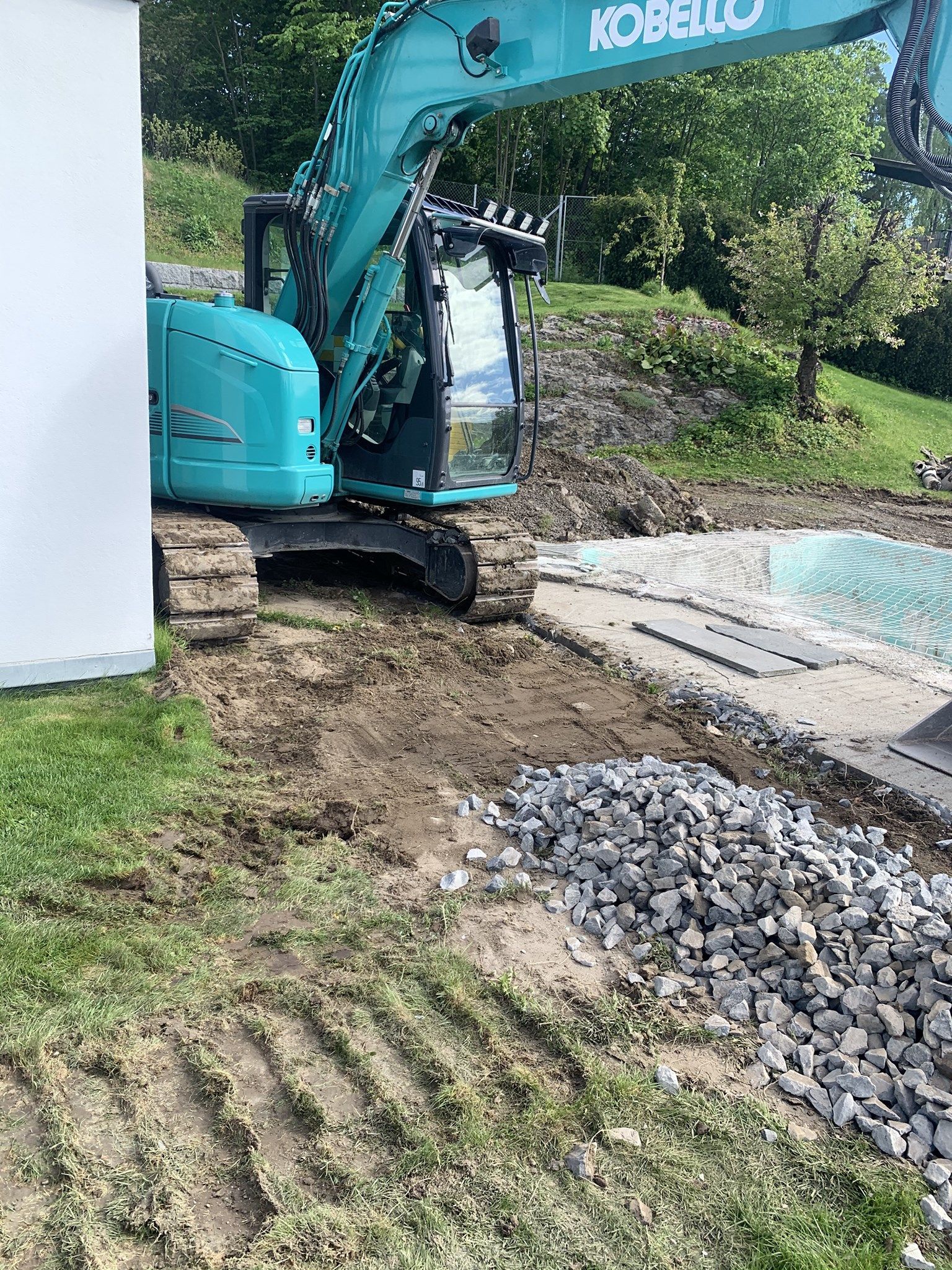 Blågrønn KOBELCO gravemaskin graver ved siden av en steinrøys på en byggeplass.