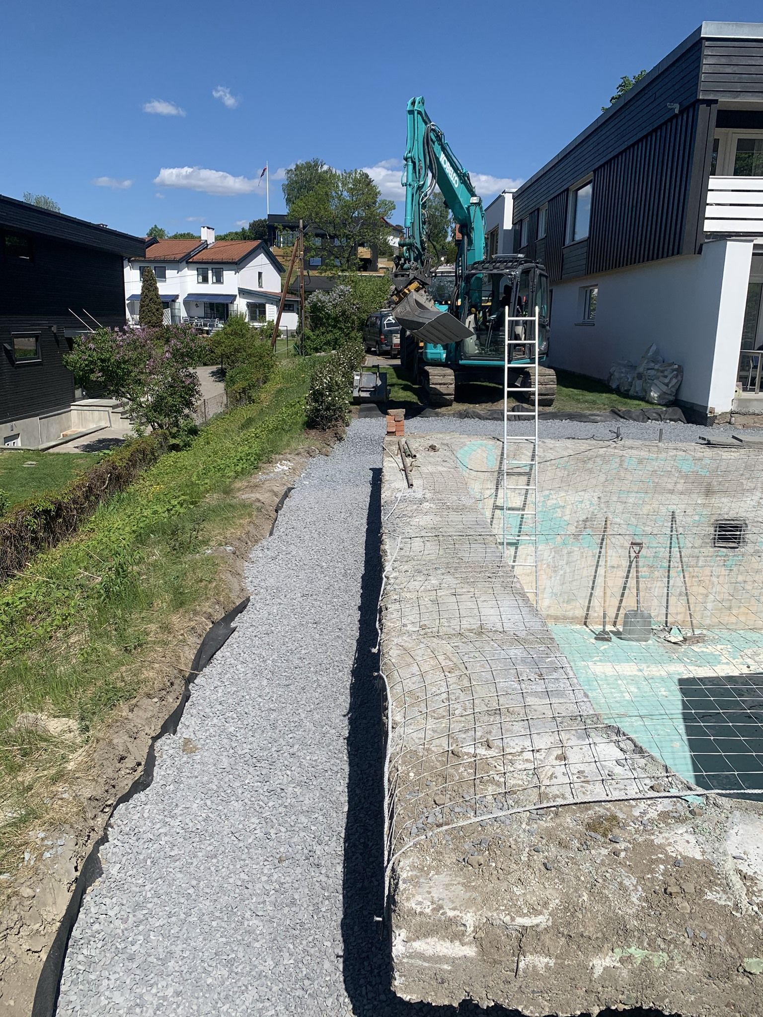 Byggeplass med gravemaskin ved siden av en grusvei og delvis revet bygning under en blå himmel