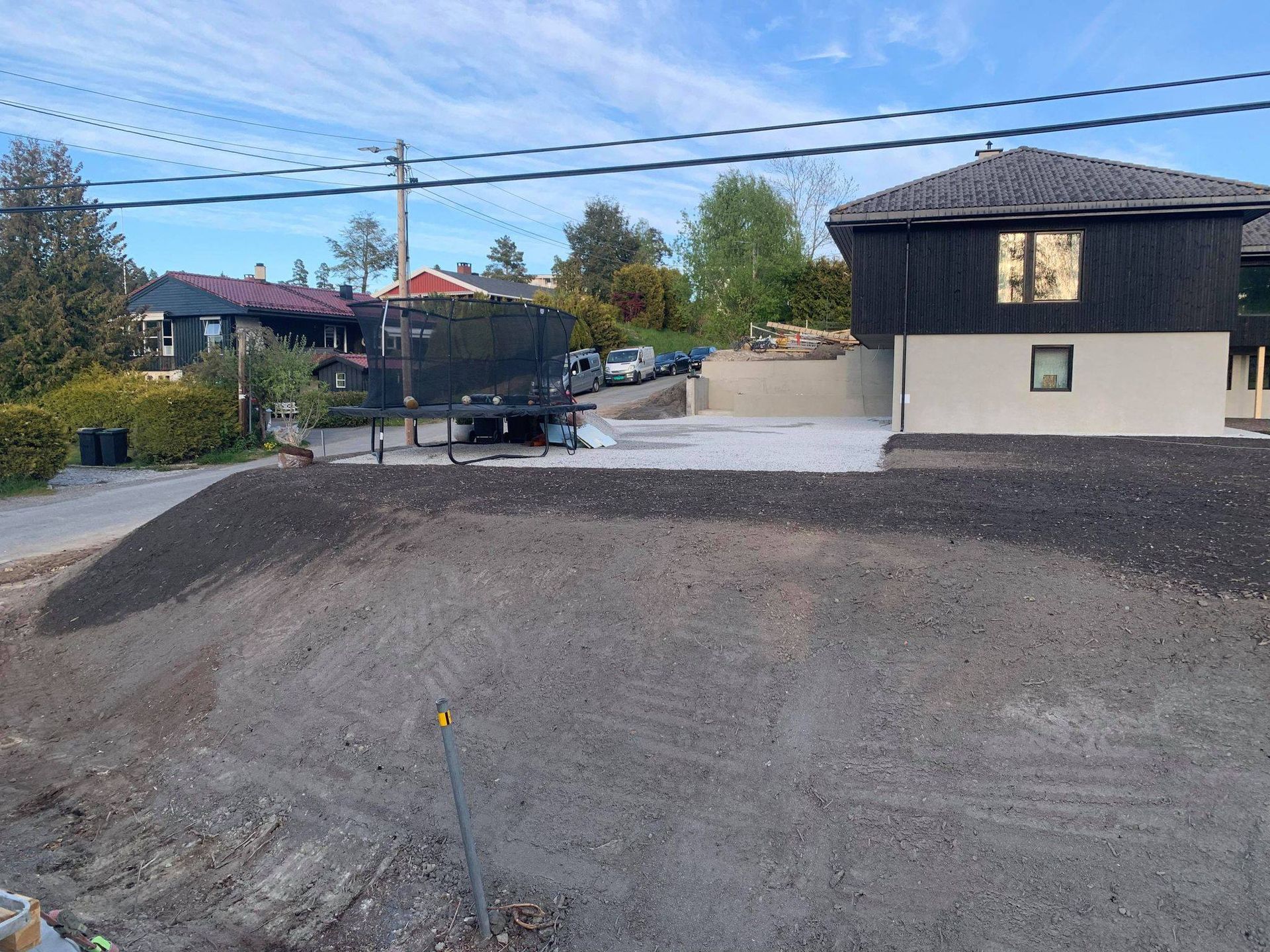 Grustomt ved siden av et hus og en lastebil, med kraftledninger og trær under en klar himmel.