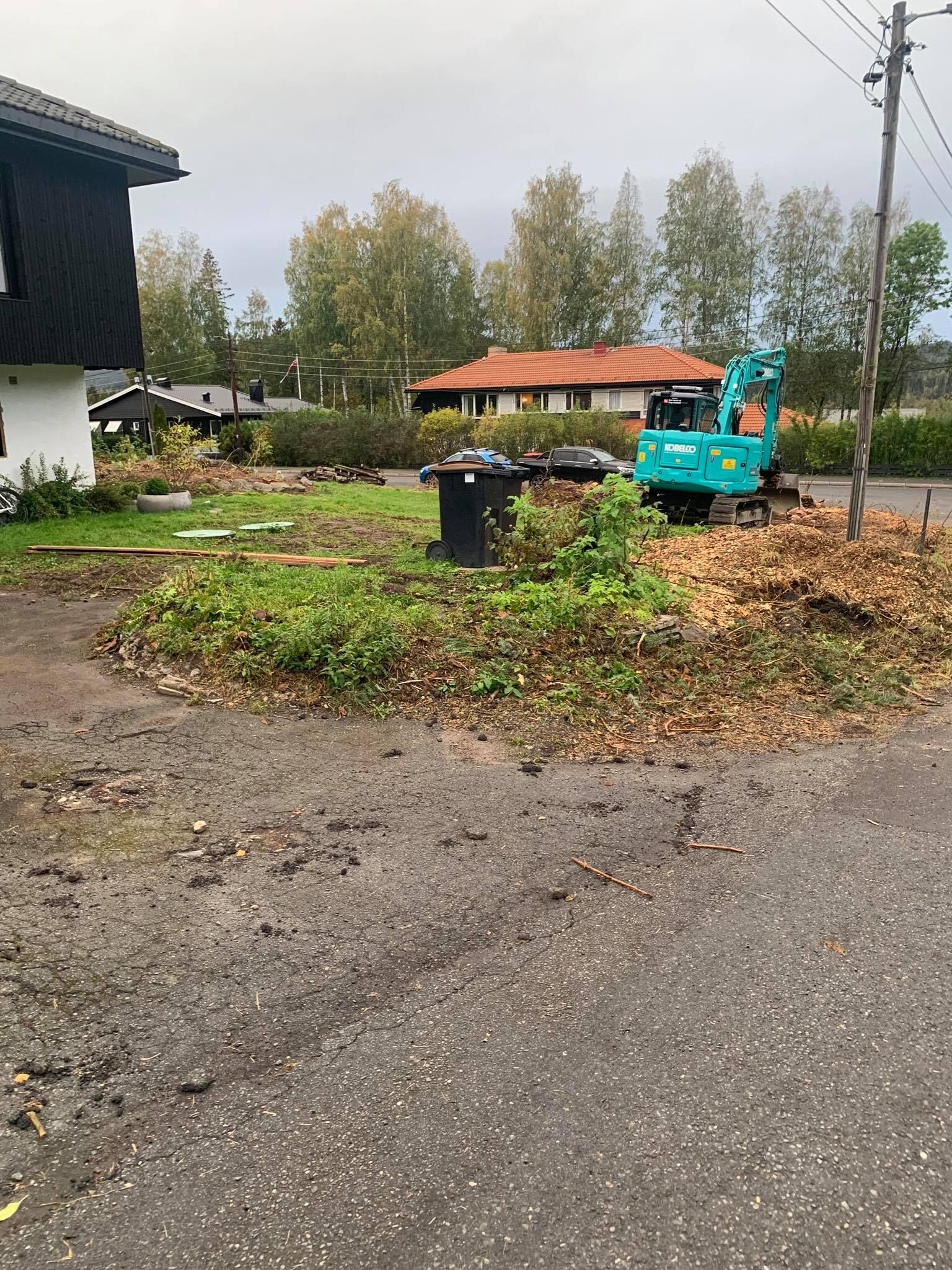 Gravemaskin rydder løv i en våt veikant ved siden av et hus og en ledningsstolpe