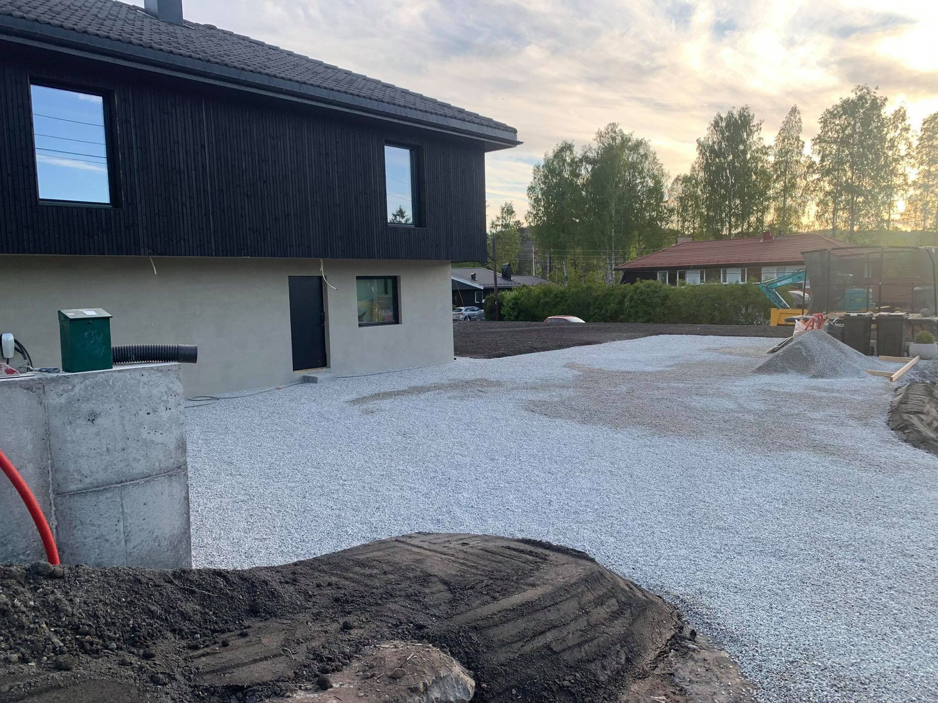 Nytt husfasade med grusoppkjørsel, uferdig jord i forgrunnen og trær ved solnedgang