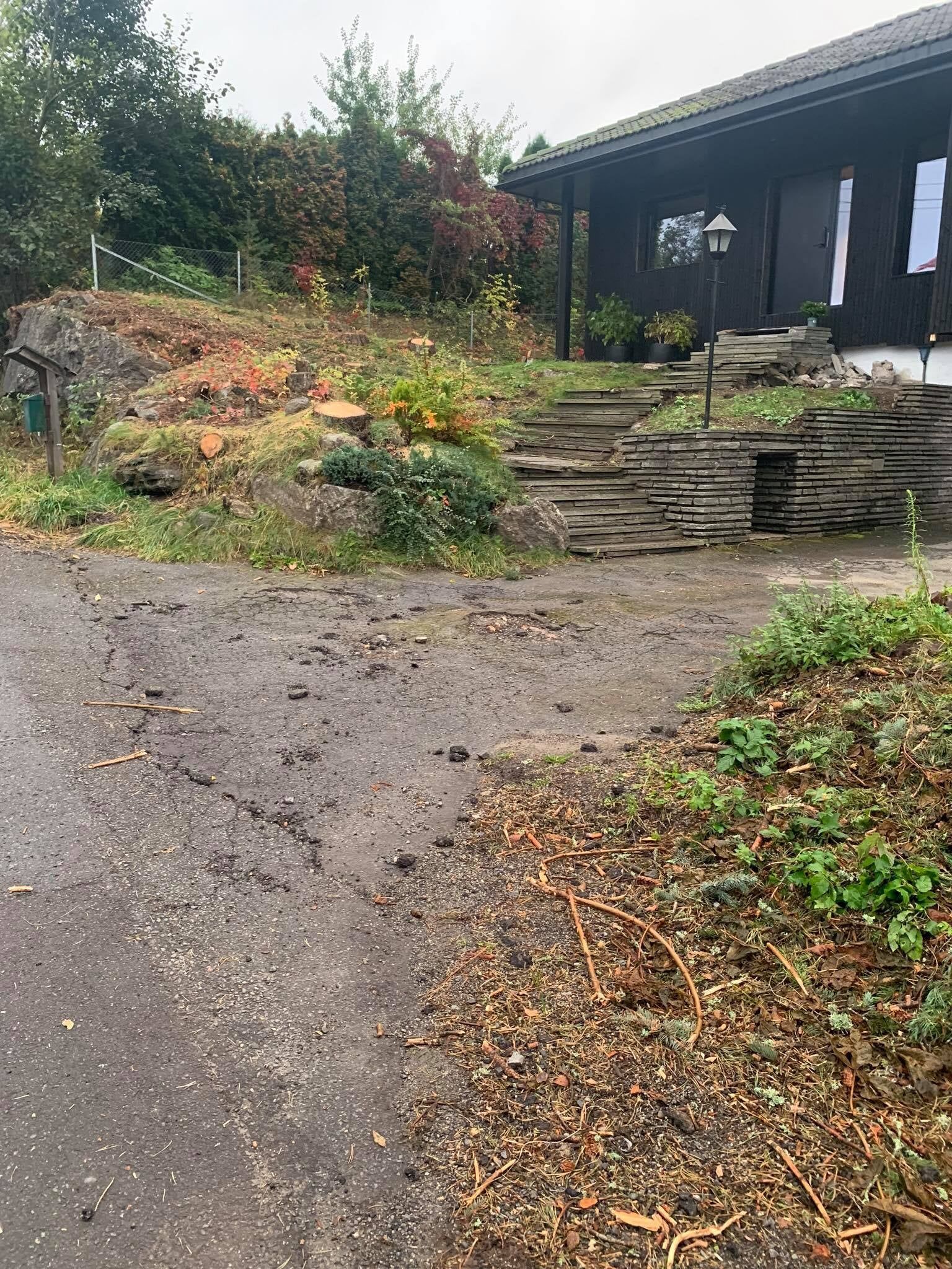 Grusvei som fører til steintrapper og et mørkt hus ved siden av en våt, løvrik hage.