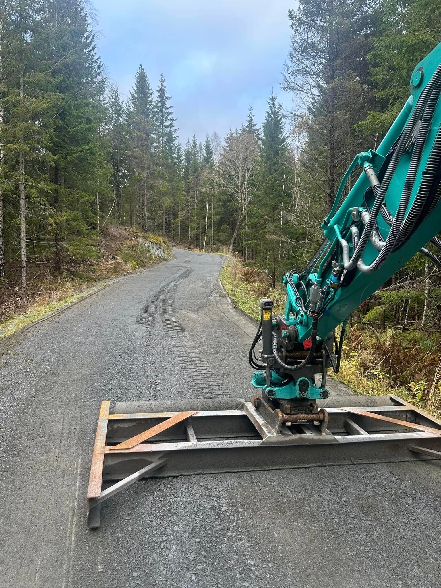 Gravemaskinbom festet til en base på en grusvei i skogen, omgitt av eviggrønne trær.