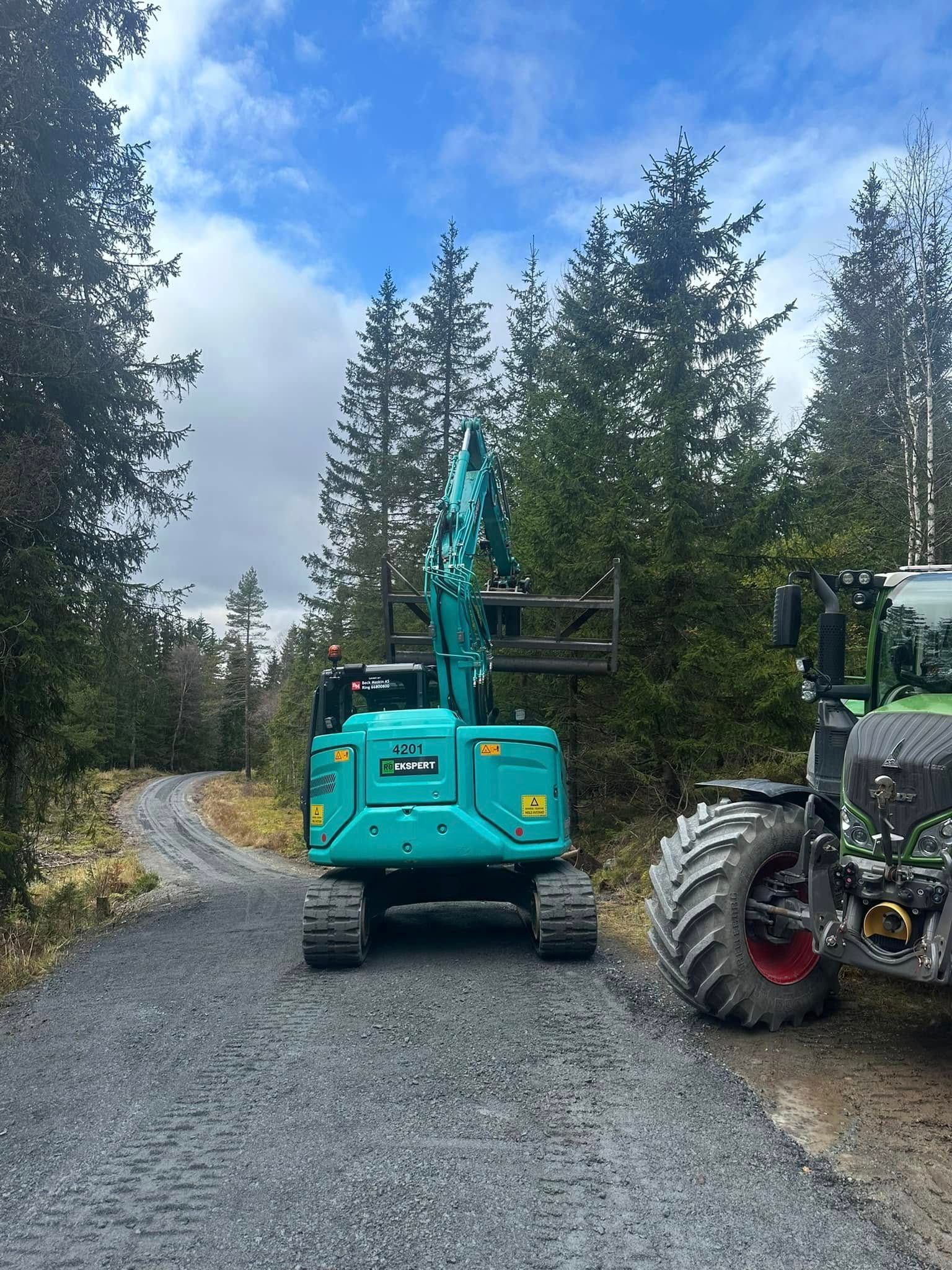 Turkis gravemaskin på en grusvei i en skog, med en traktor delvis synlig til høyre.