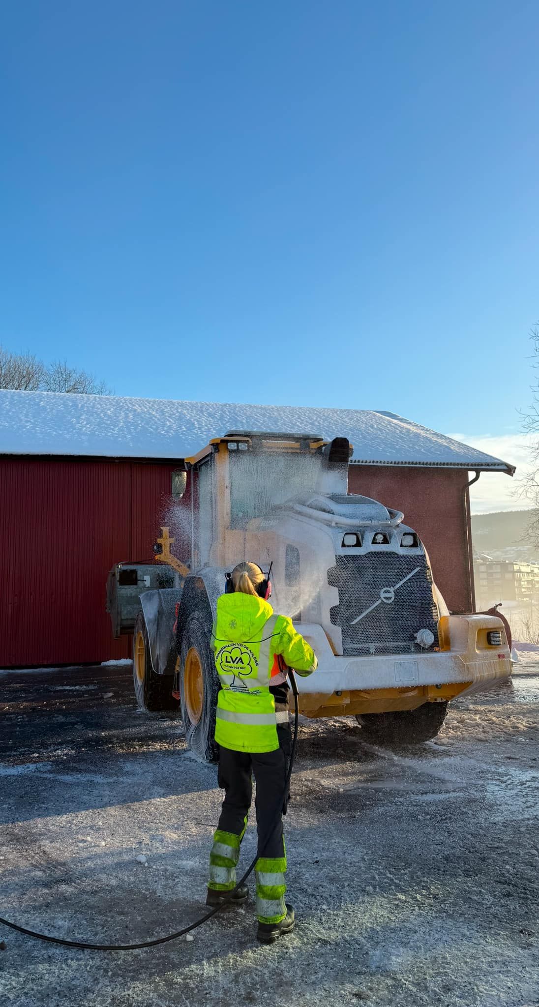 Arbeider i neonsikkerhetsutstyr sprayer skum på en stor maskin utenfor en rød bygning.