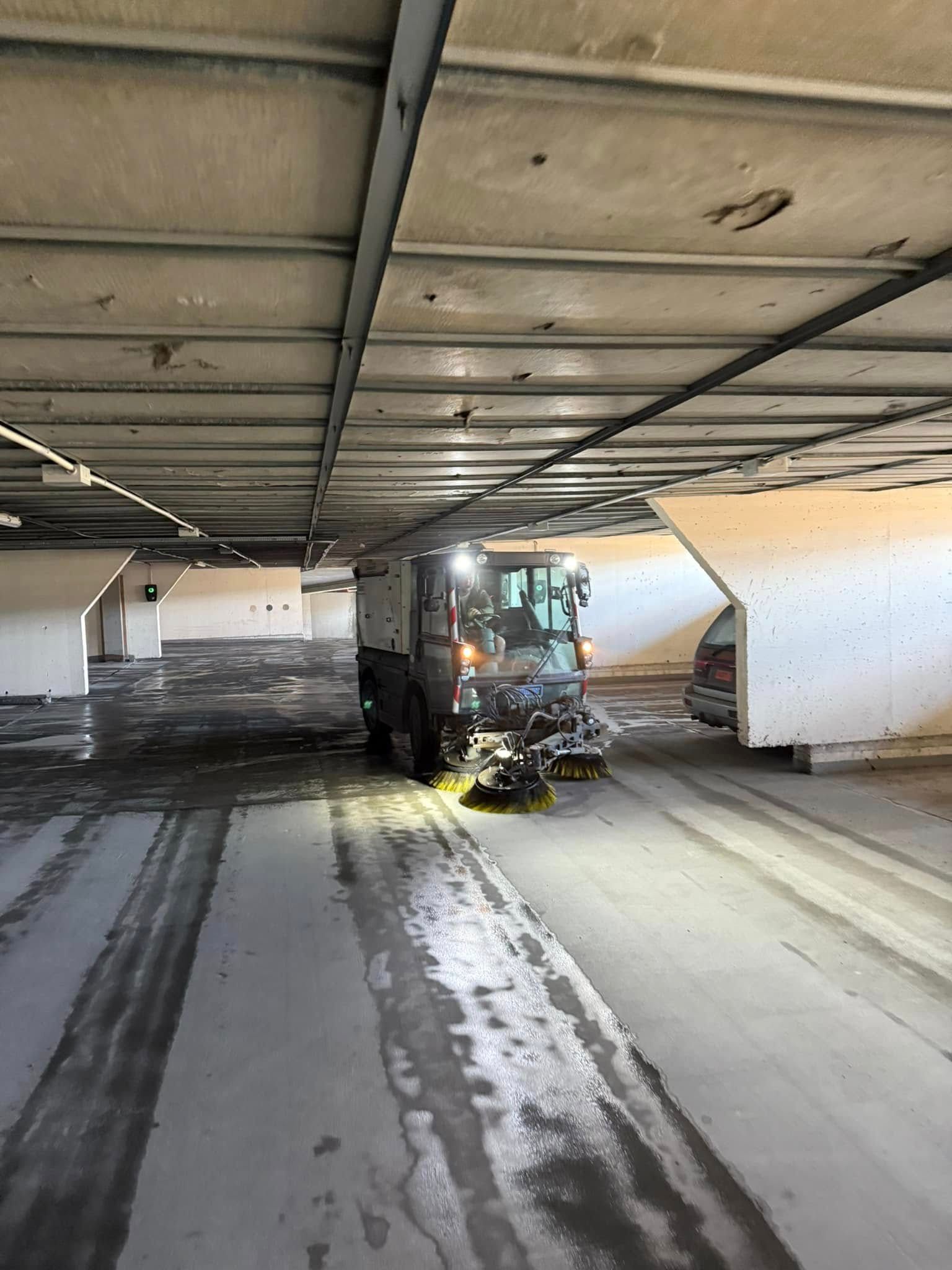 Tomt parkeringshus med en midtsøyle, overliggende bjelker og mørke dekkmerker på betonggulvet.