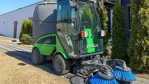 Grønn gatefeier parkert ved en bygning, med blå og svarte børster synlige foran.