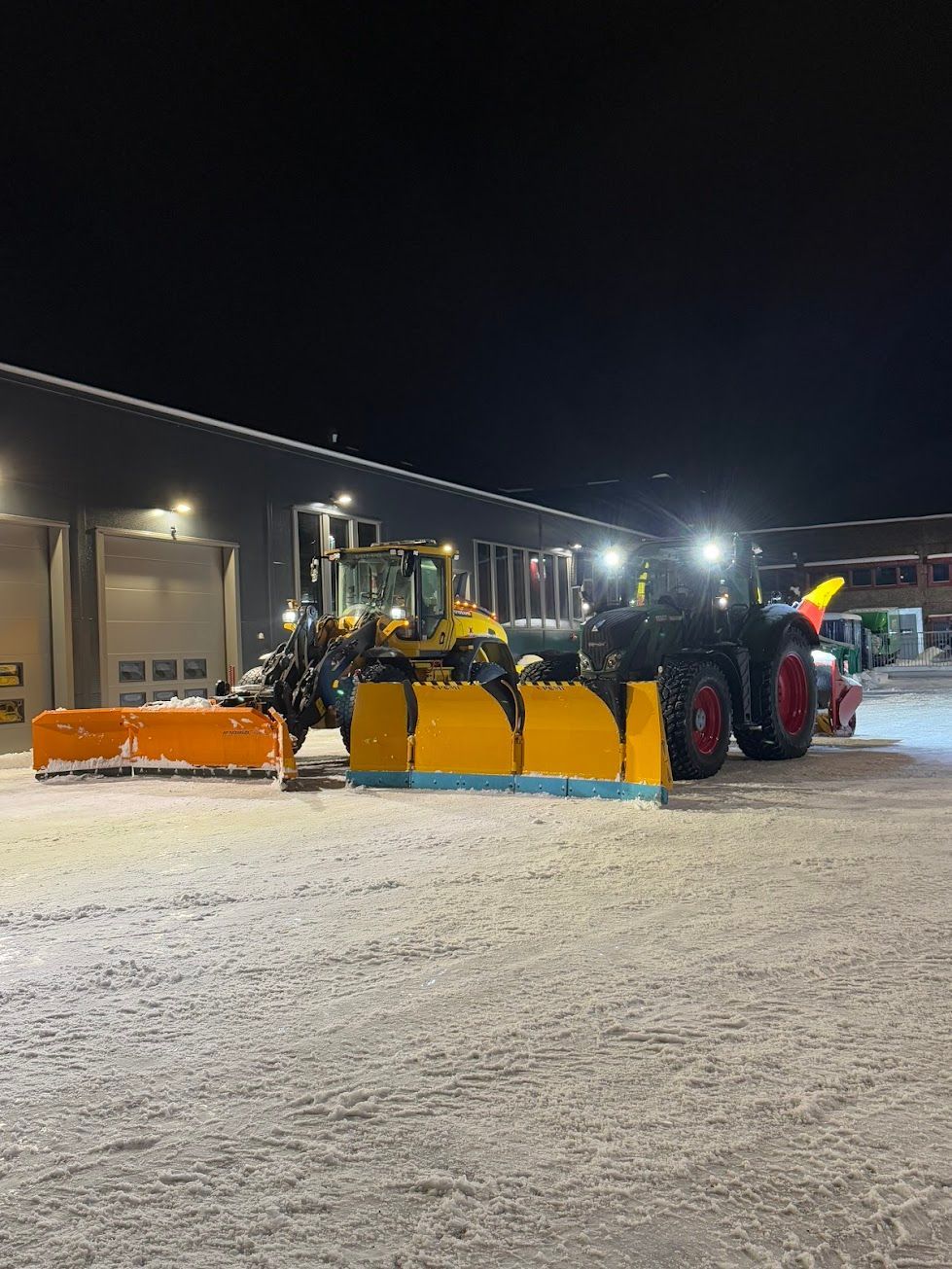 En blågrønn gravemaskin laster en blå dumper med jord. Sett mot en lys blå himmel og spredte grønne trær.