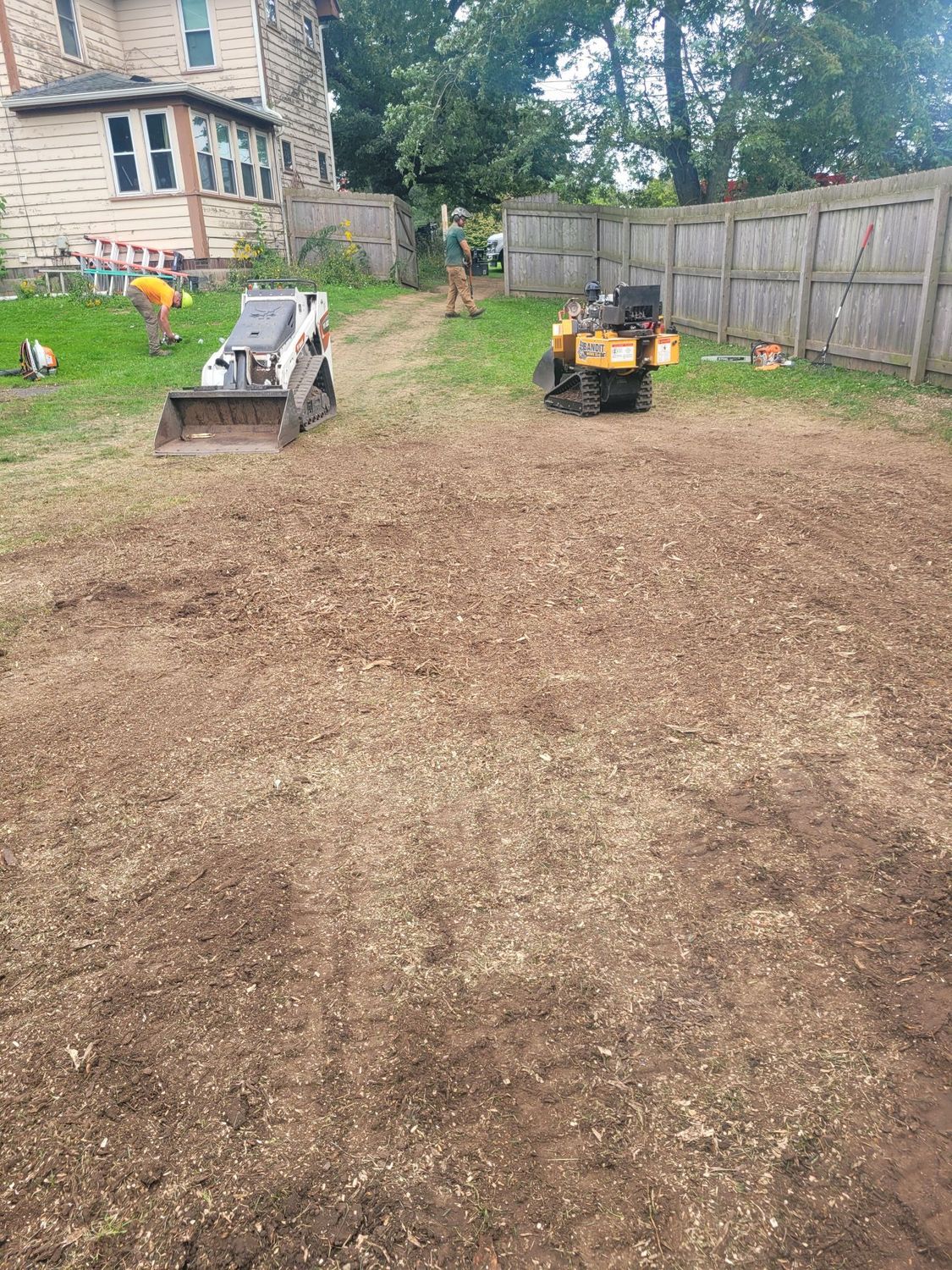 A construction crew operates a skid steer and stump grinder in a yard with wood chips covering the ground near a house.