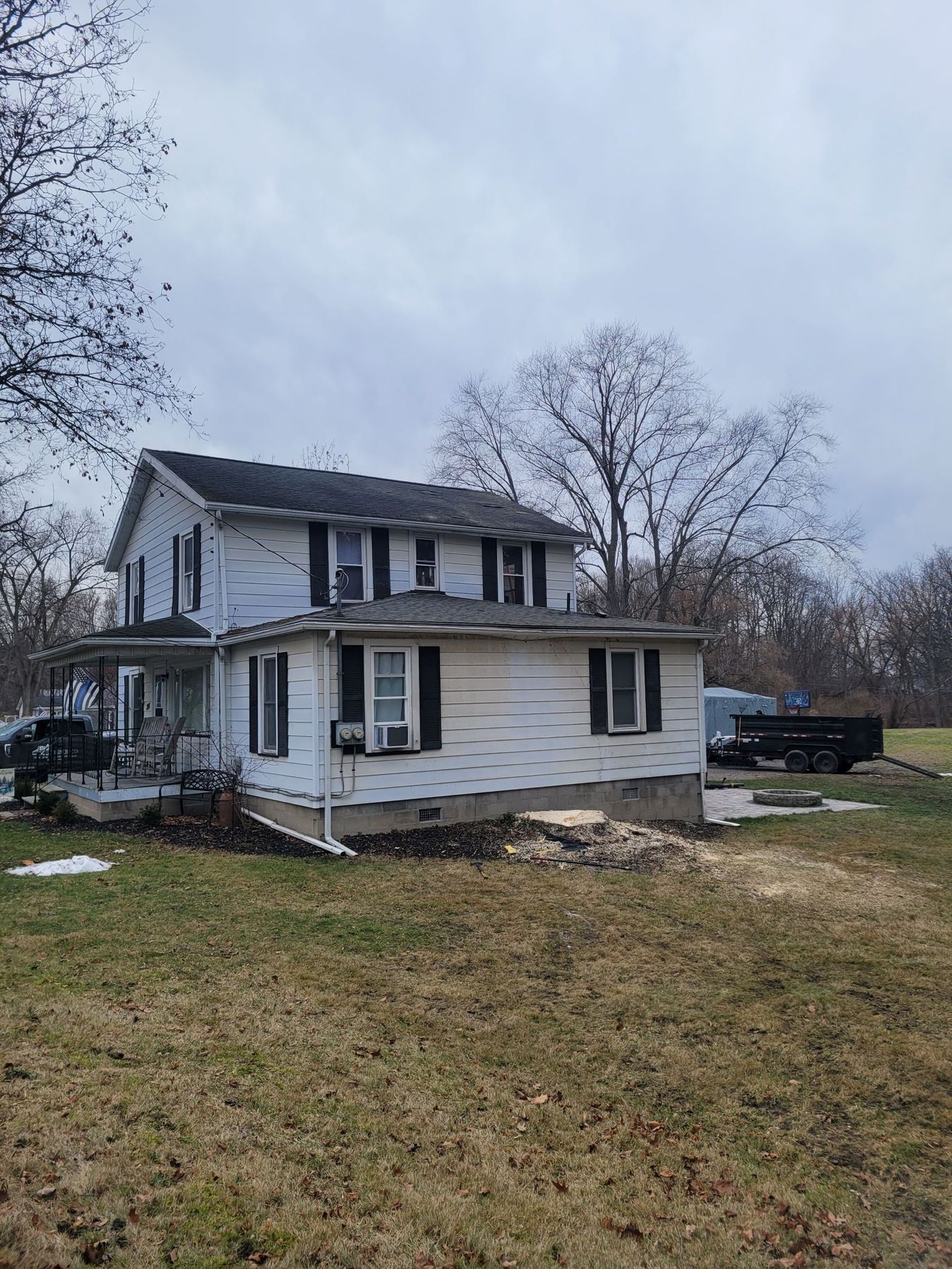 A white, two-story house with black shutters and a front porch, set against a cloudy sky with a trailer in the yard.