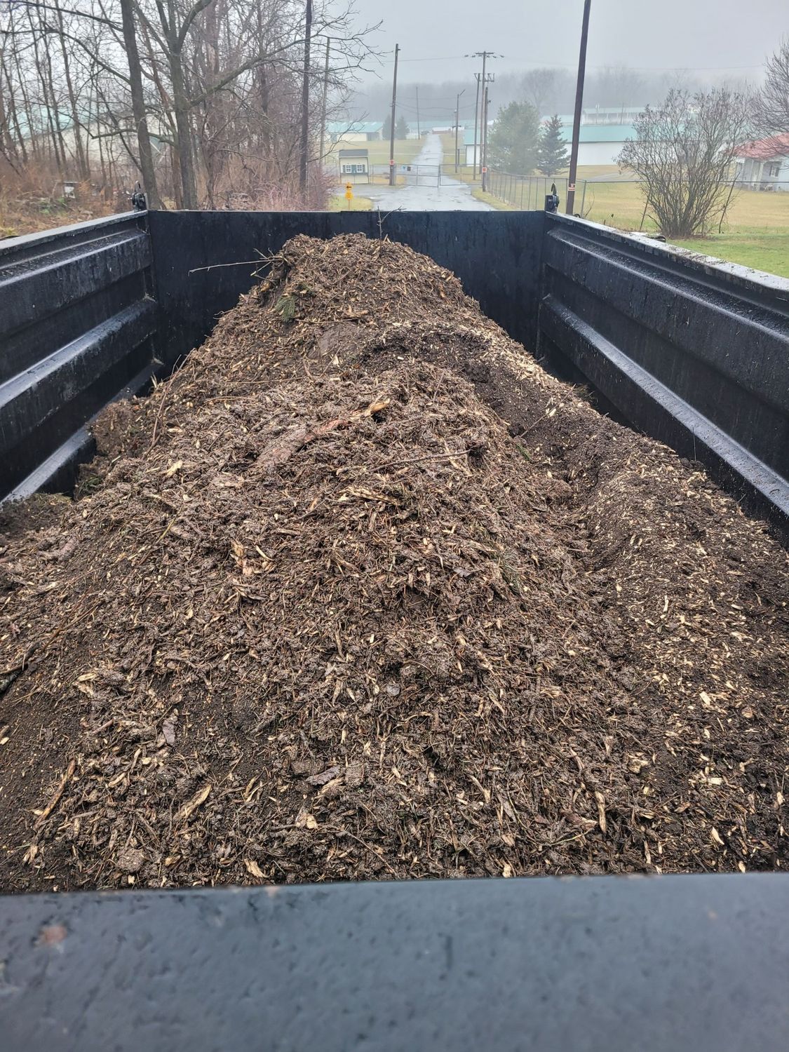 A full dump truck bed loaded with a large pile of dark, shredded wood mulch against a rural, misty background.