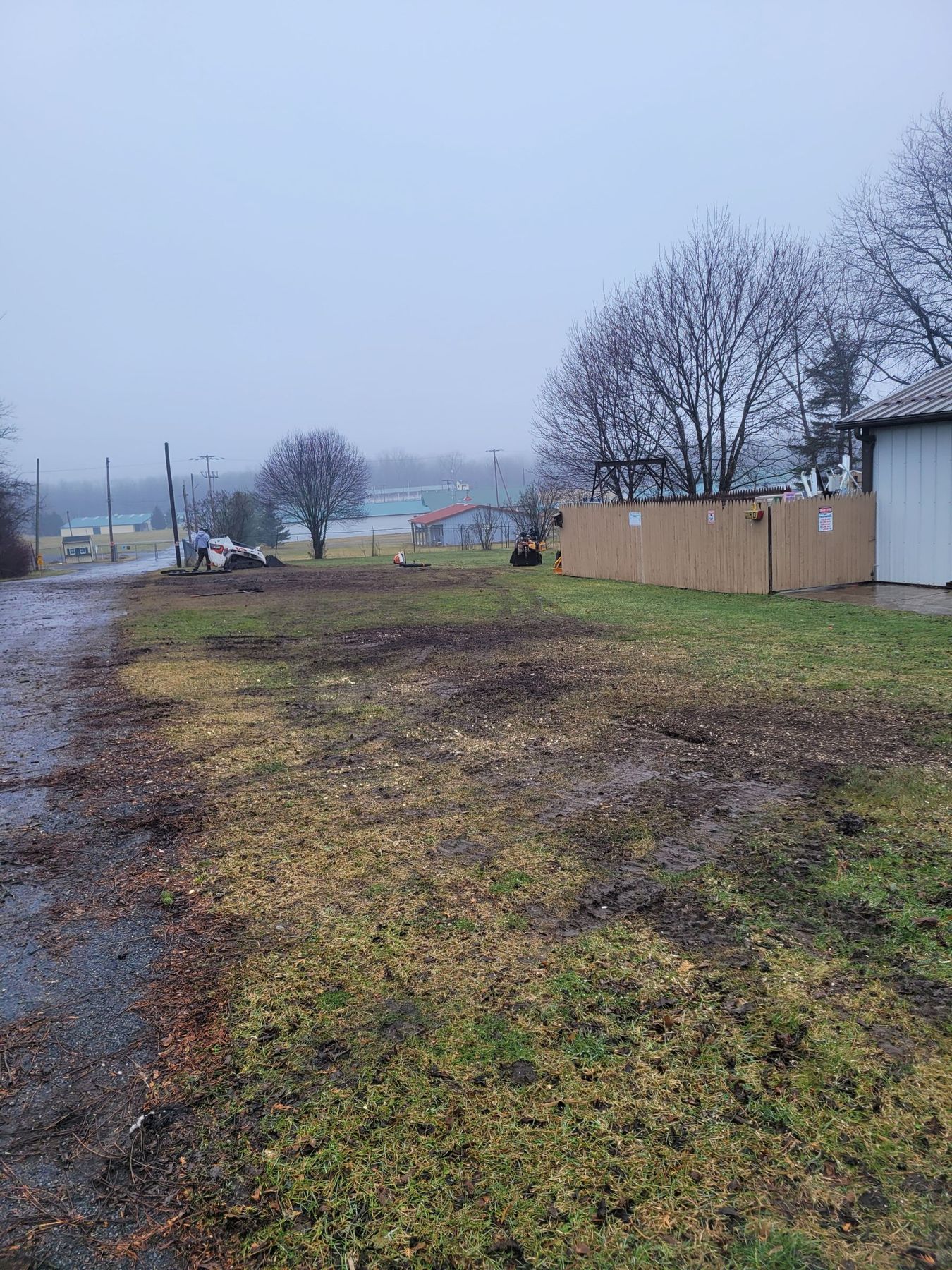 A muddy, overcast field with a wooden fence, bare trees, and a small building under a grey sky.