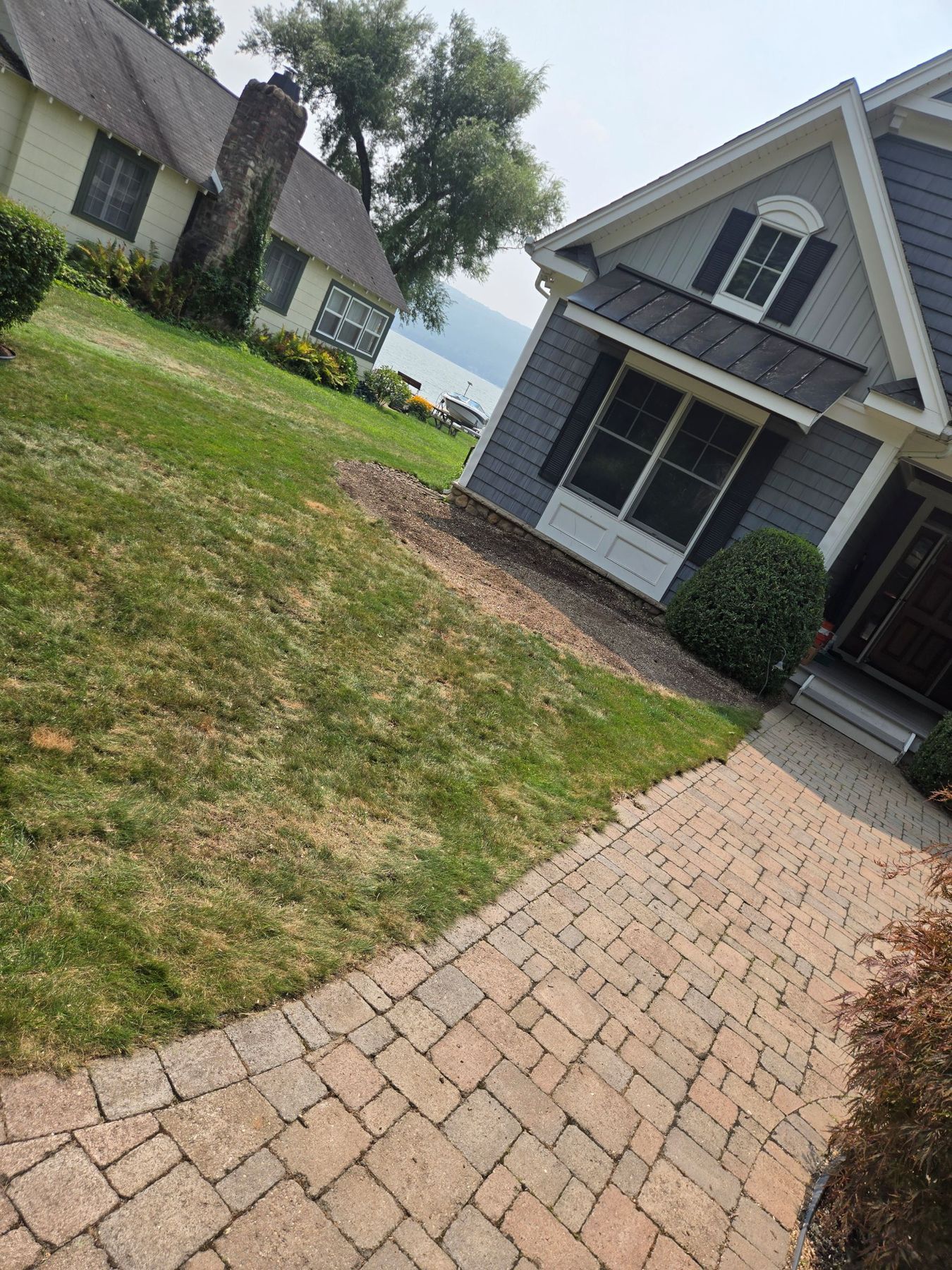 A paved brick walkway leads toward the blue-gray siding and windows of a suburban house on a sunny day.