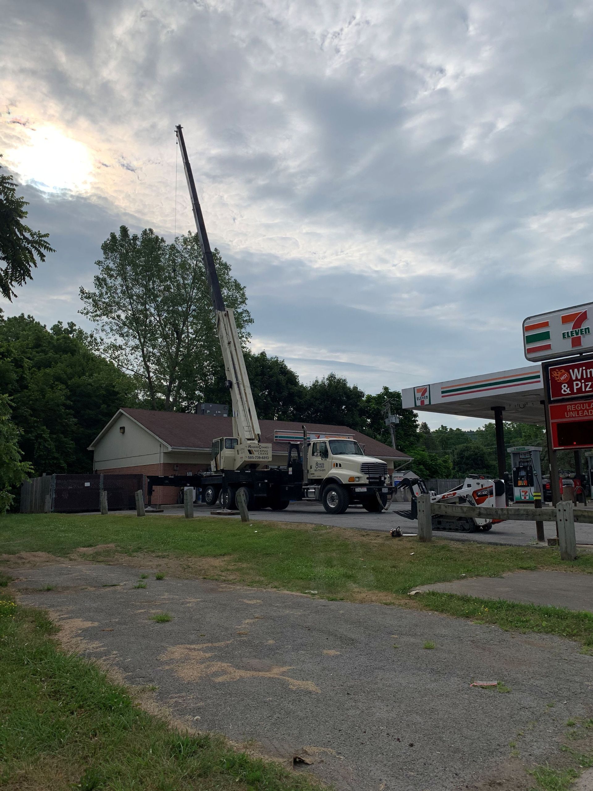 A white boom truck with an extended crane parked at a 7-Eleven gas station under a cloudy, overcast sky.