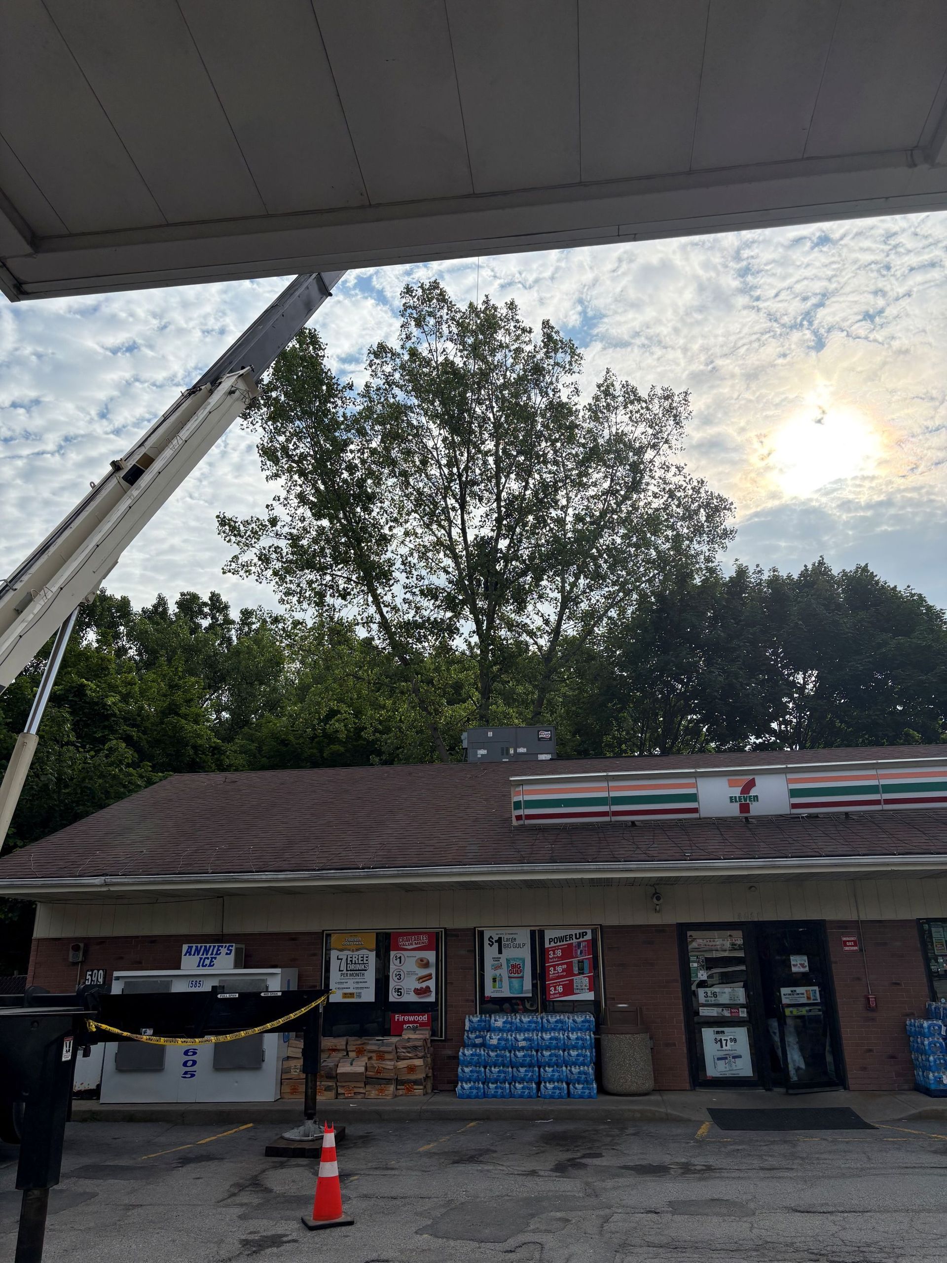 A 7-Eleven store exterior under a blue sky with a cherry picker crane extended toward trees behind the building.