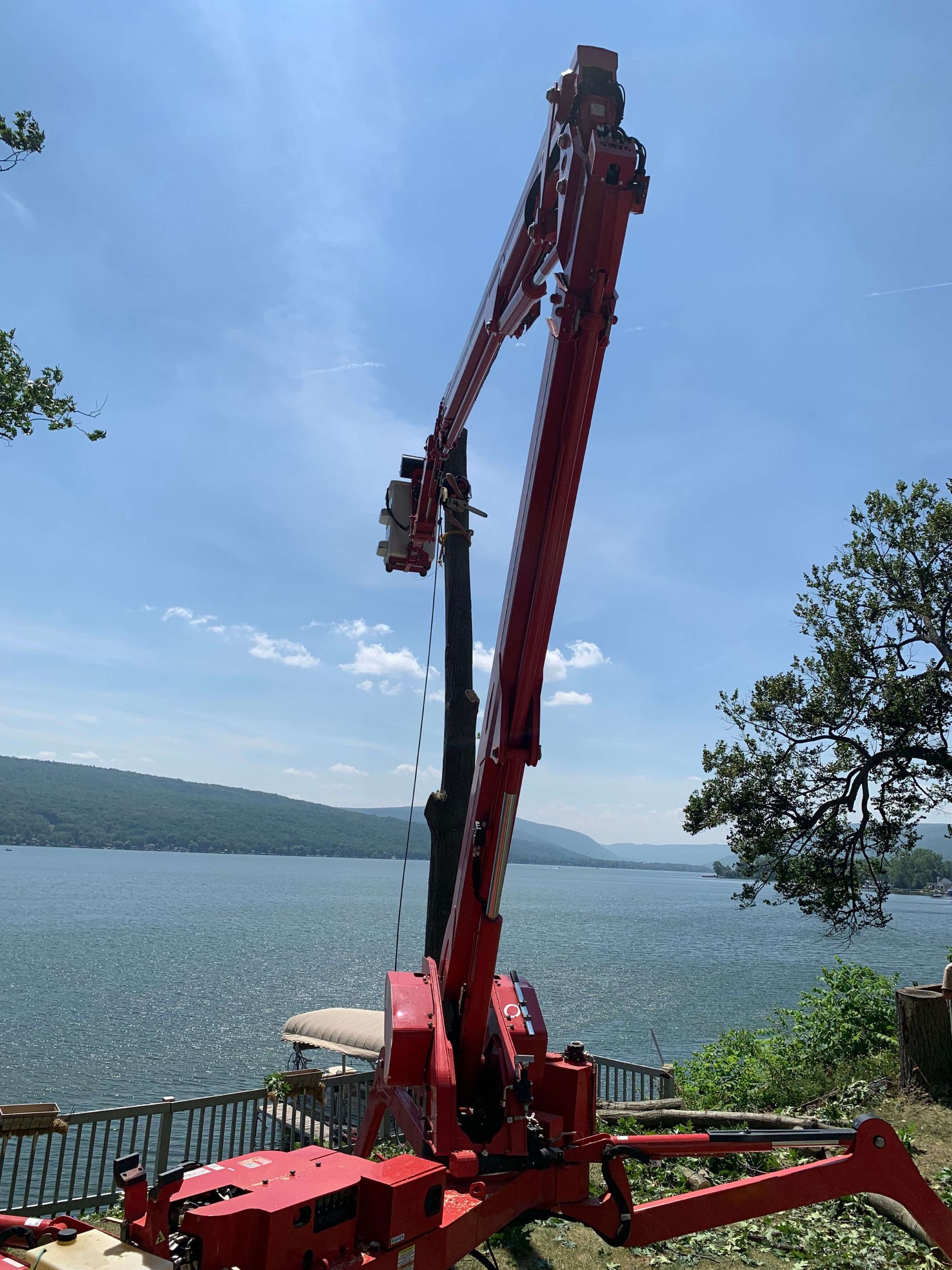 A red boom lift extends its arm over a scenic lake, with outriggers deployed on a grassy slope under a bright blue sky.