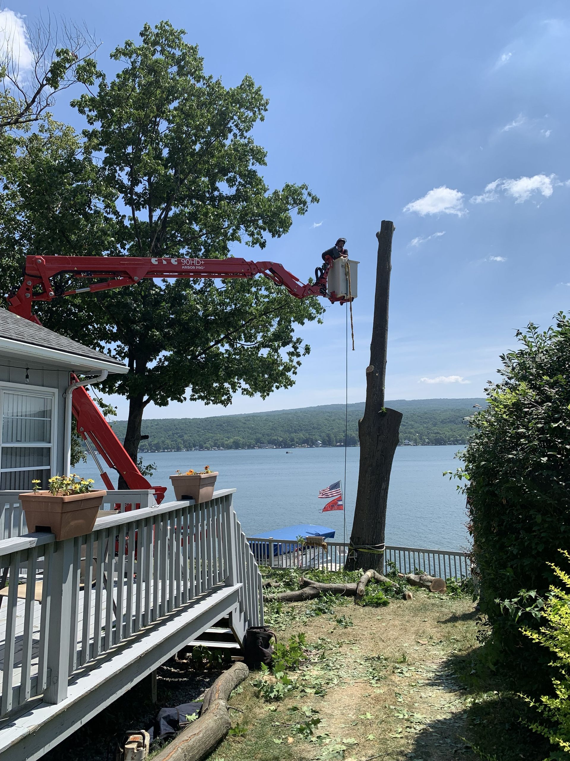 A person in an aerial lift bucket trims a tall, bare tree trunk by a lake near a house deck.