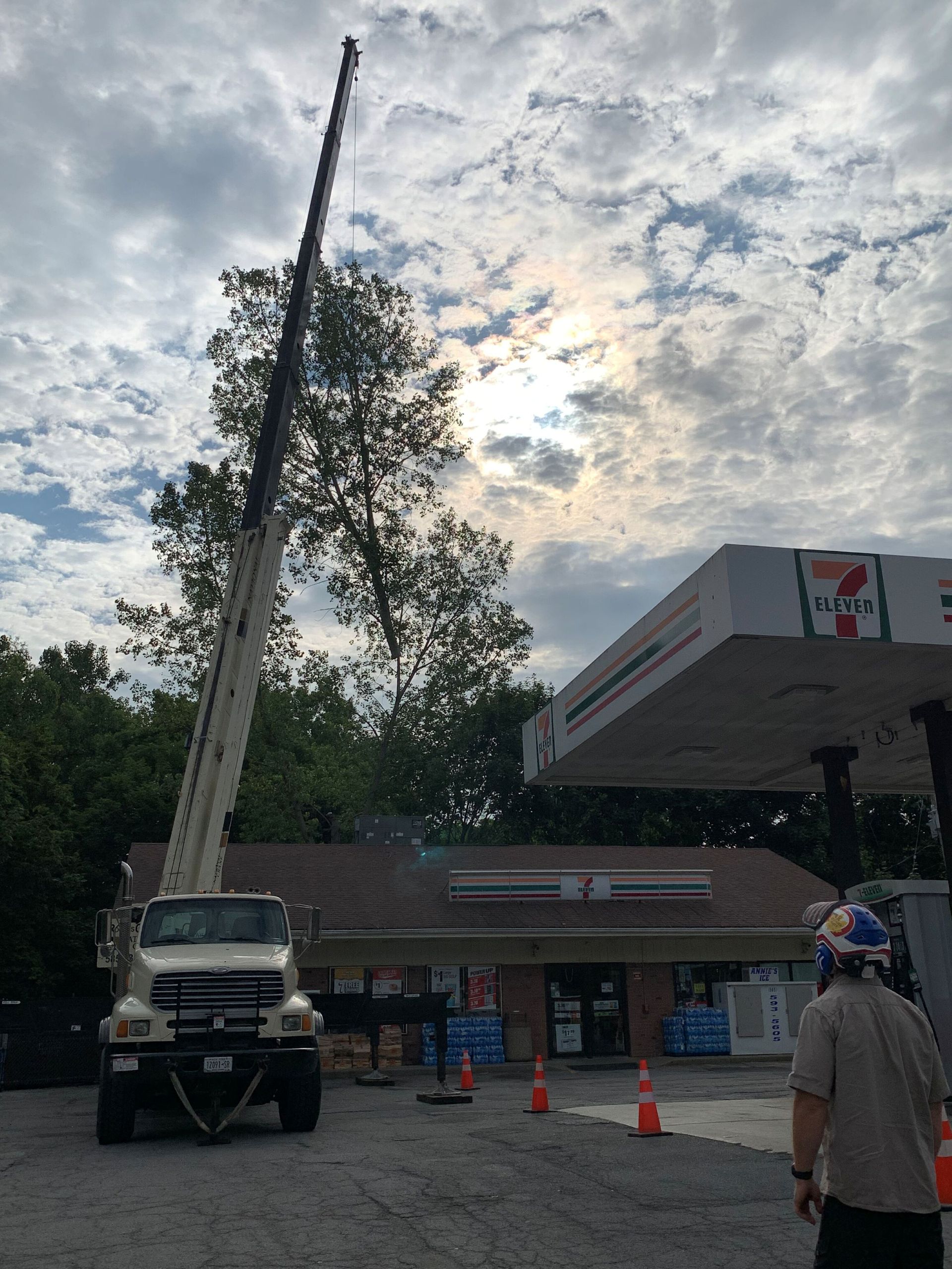 A crane is parked at a 7-Eleven gas station, with its boom extended high into the sky near a tree.
