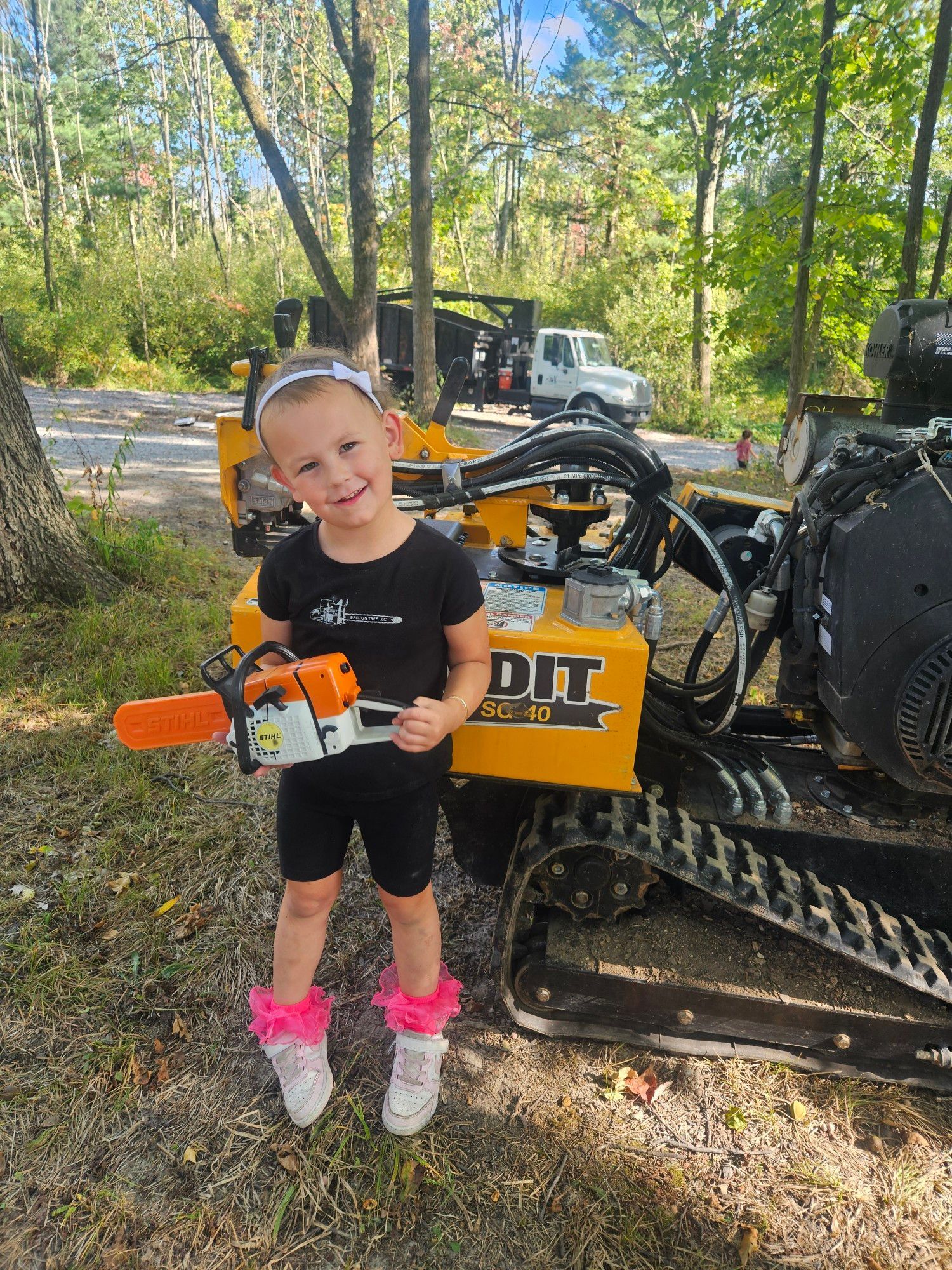A person stands in a wooded area holding a small toy chainsaw in front of a yellow Bandit wood chipper.